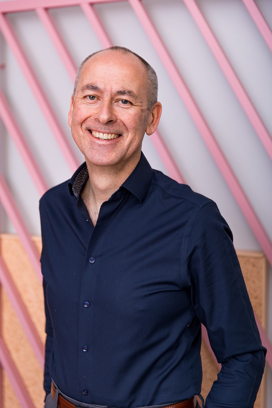 A smiling middle-aged man with a bald head, wearing a navy blue button-up shirt, standing in front of a wall with pink diagonal beams.