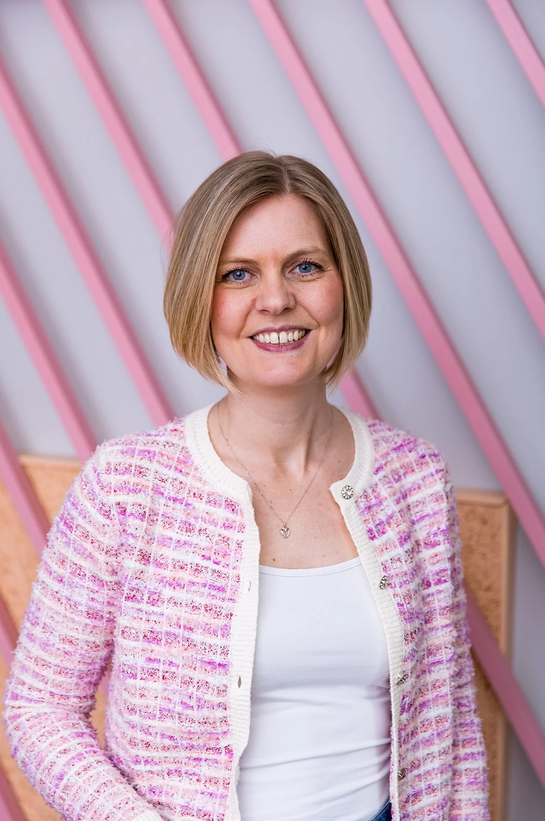 A smiling woman with shoulder-length blonde hair, wearing a pink and white textured jacket and a white top, standing in front of a background with pink diagonal beams.