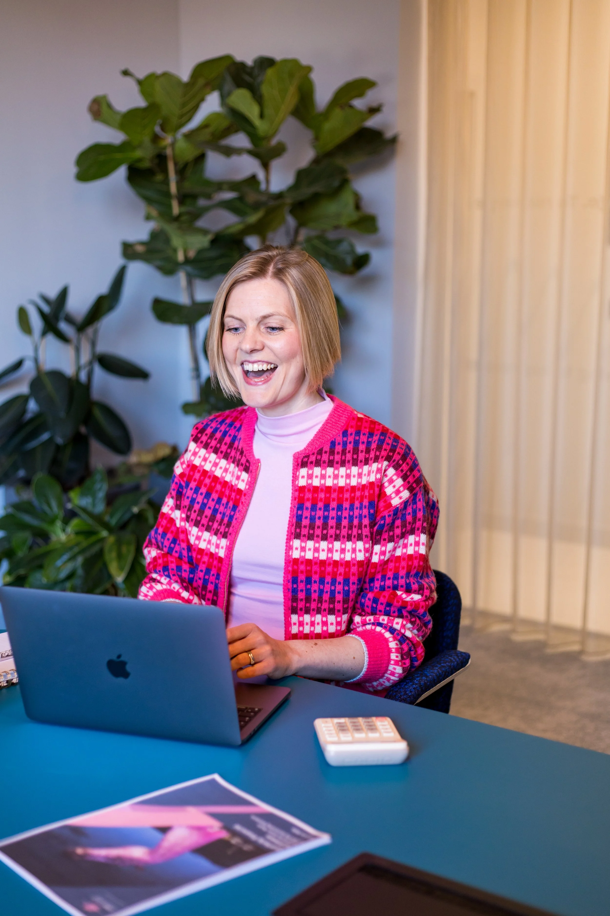 Woman with light brown hair in a bob cut, smiling and wearing a pink and purple patterned sweater, sitting at a blue table with a silver MacBook, a magazine, and a small keyboard. There are large green houseplants and light-colored wooden panels in the background.