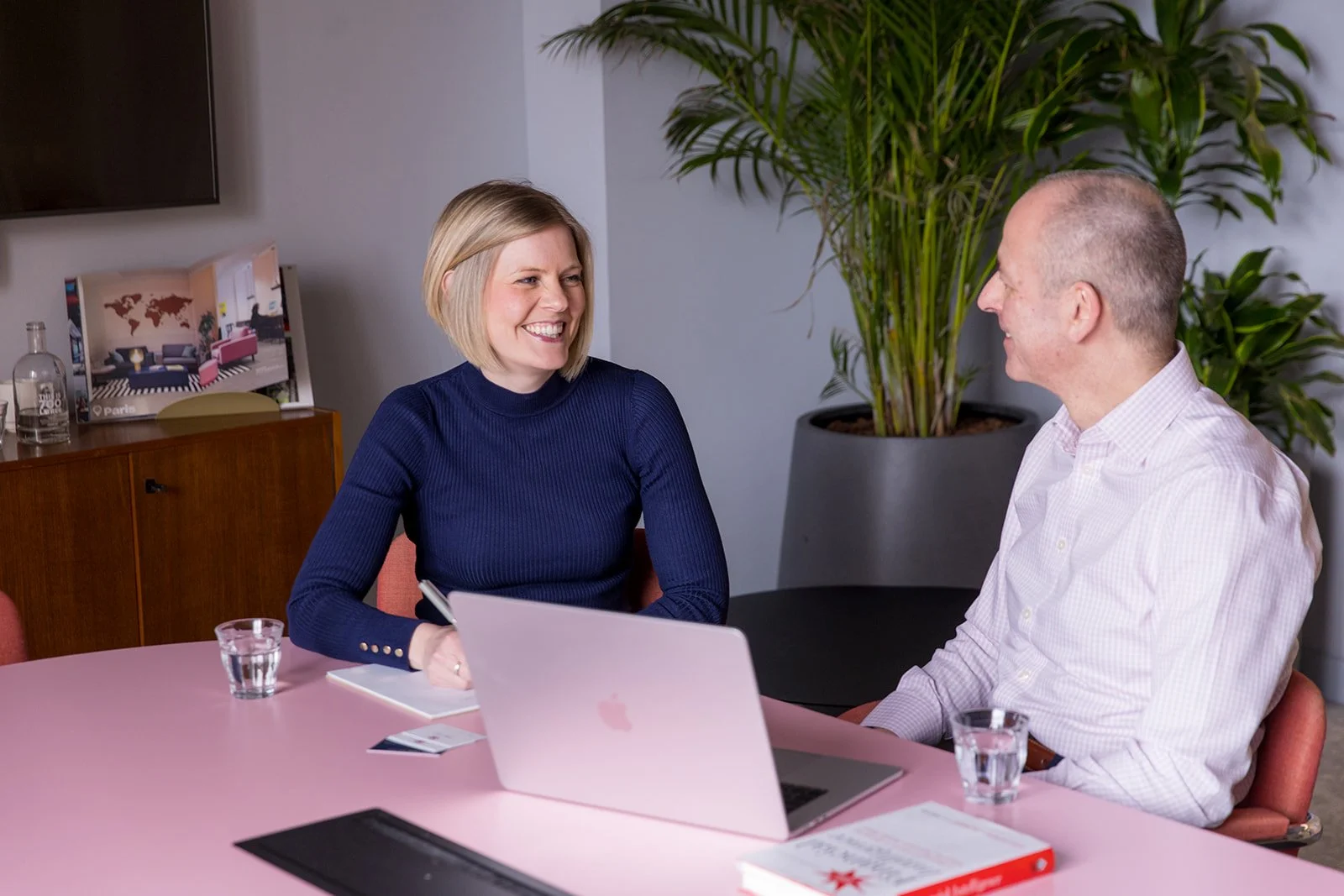 A woman and a man sitting at a pink table in a modern office space, engaged in conversation. The woman has blonde hair and is wearing a navy blue sweater, smiling. The man is bald, wearing a light pink checkered shirt, and smiling back. There is an open laptop, glasses of water, and some documents on the table. In the background, there is a large green plant and a cabinet with a framed picture of a room, a glass bottle, and a world map on the wall.