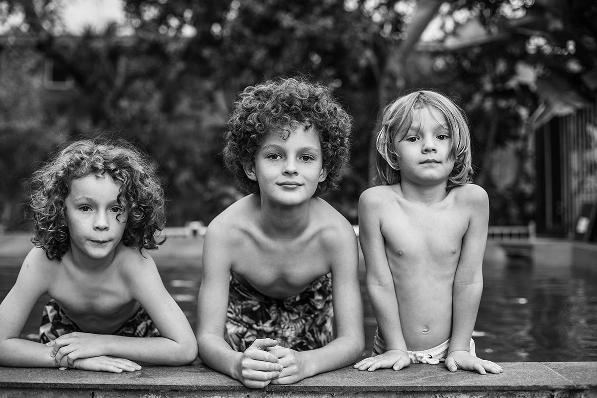 Three children with curly hair leaning on a pool edge, wearing swim trunks, in black and white.
