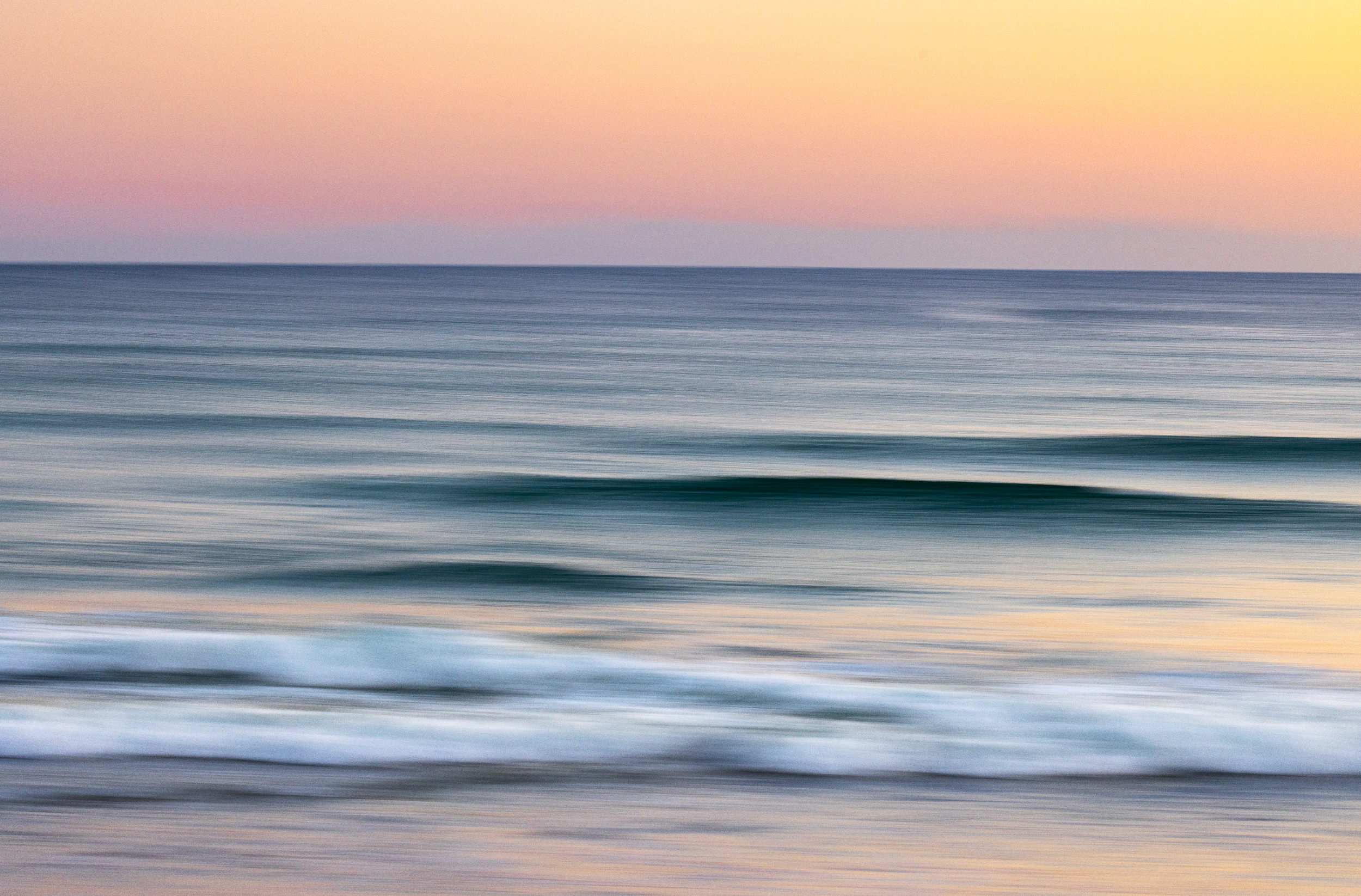 Blurred image of ocean with gentle waves during sunset, pastel pink and orange sky.