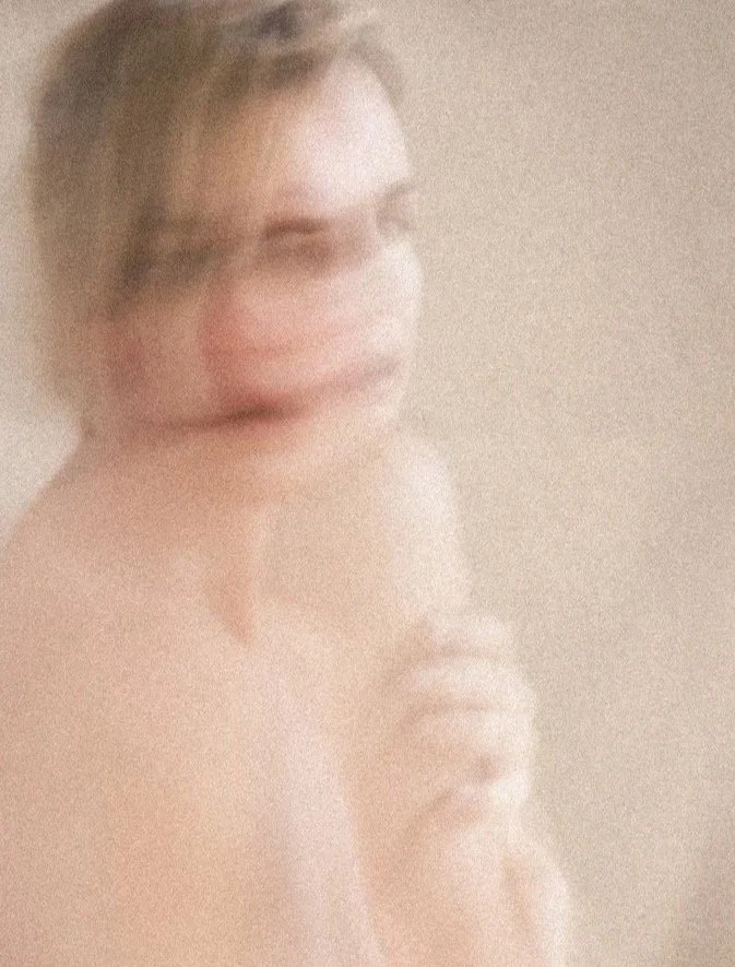 A blurred, artistic photo of a woman with light skin, short hair, and a subtle smile, posed with her hands near her chin, against a neutral background.