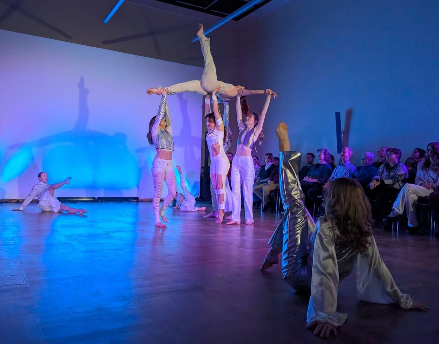 Tänzer in einem Bühnenauftritt, einige in akrobatischen Posen, während andere im Publikum zuschauen.