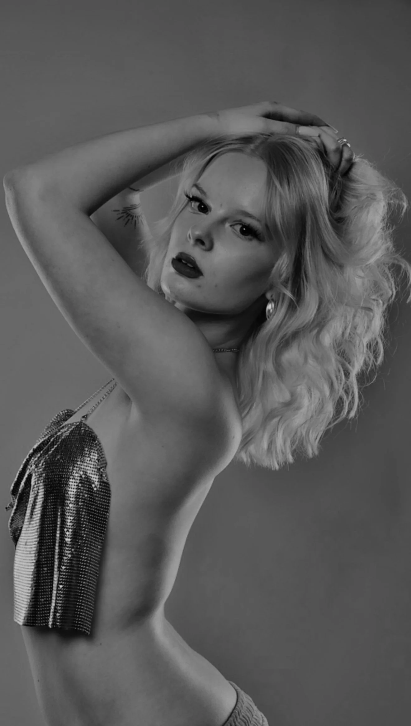 Schwarz-weiß-Fotografie eines jungen, groß gewachsenen Mädchens mit lockigem, blondem Haar, das den Oberkörper frei hat und einen Arm über den Kopf legt, während sie in die Kamera schaut.