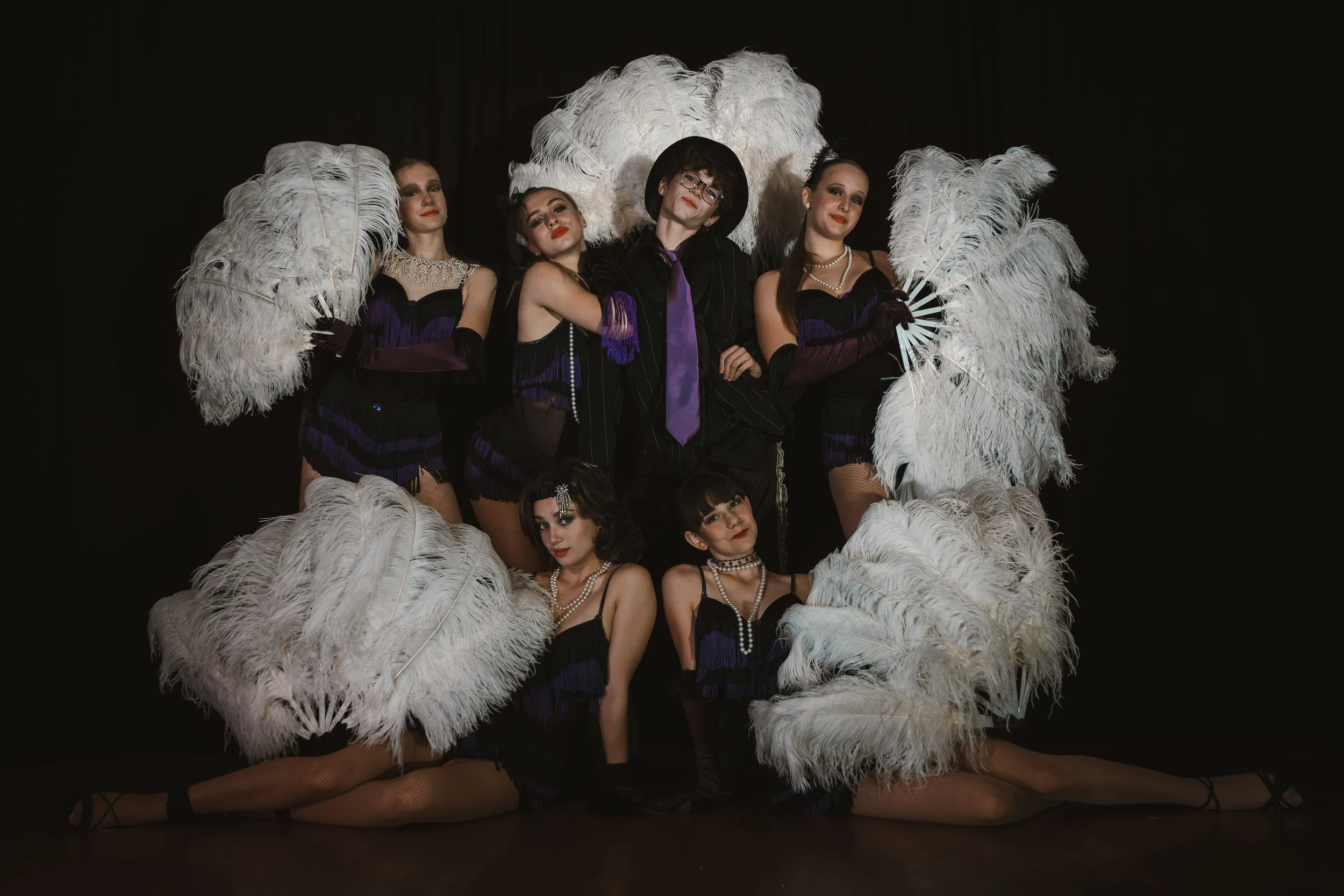 Gruppe von sieben Frauen in Vintage-Gala-Kostümen mit Federaccessoires vor schwarzem Hintergrund.