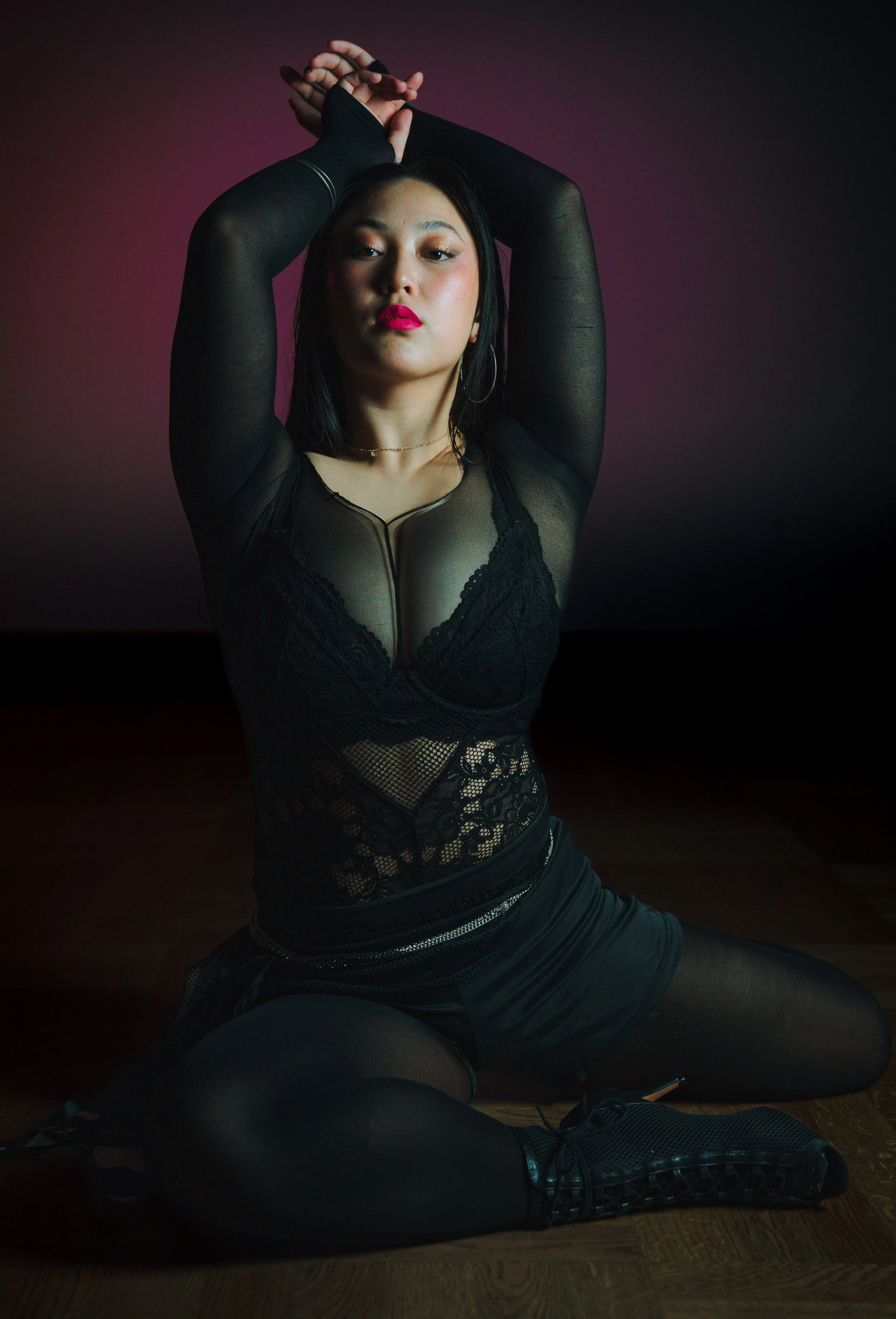 Frau in schwarzer Lingerie und Strumpfhosen sitzt kniend auf Holzfußboden vor dunklem Hintergrund mit rosa Beleuchtung, Arme über Kopf verschränkt.