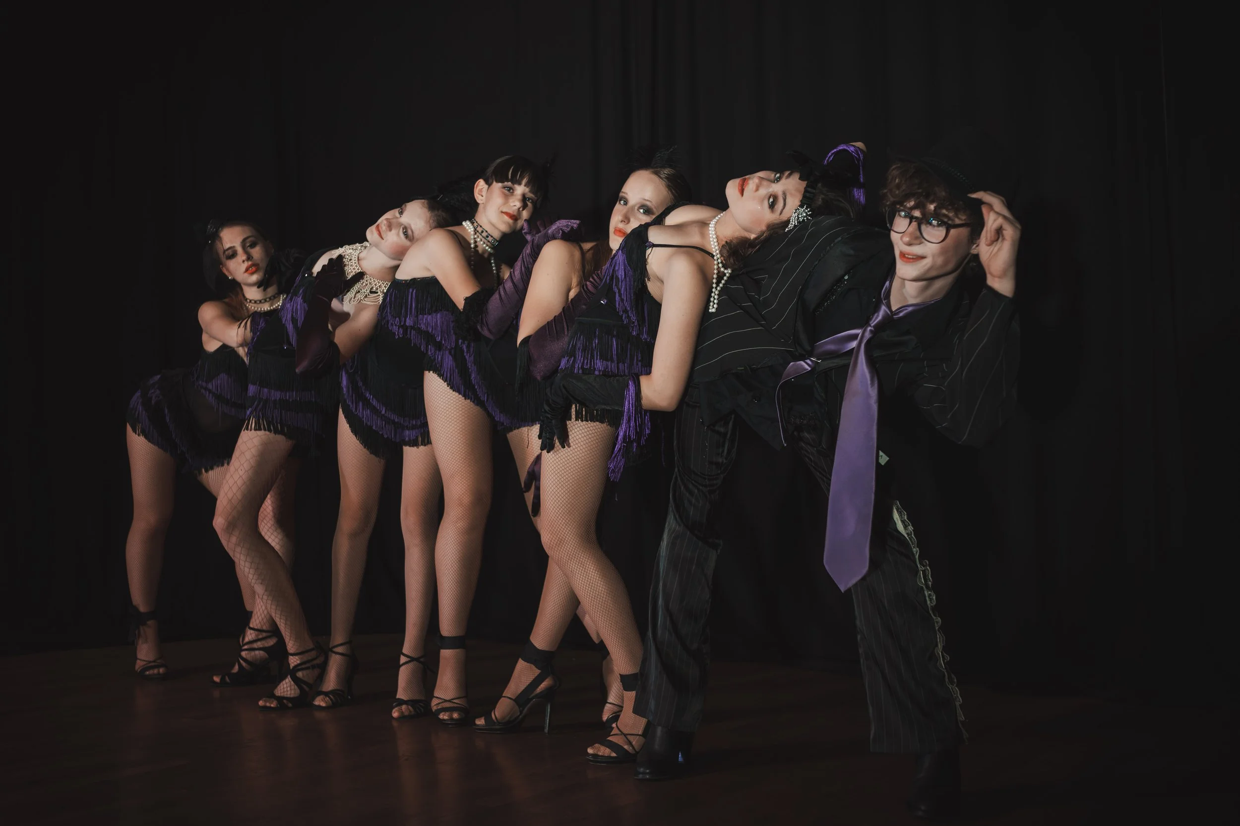 Eine Gruppe von fünf Frauen und einem Mann in Vintage-Kleidung posieren vor schwarzem Hintergrund, im Stil der 1920er Jahre, mit Flapper-Kleidern, Fischernetzen, Fransen und Hüten.