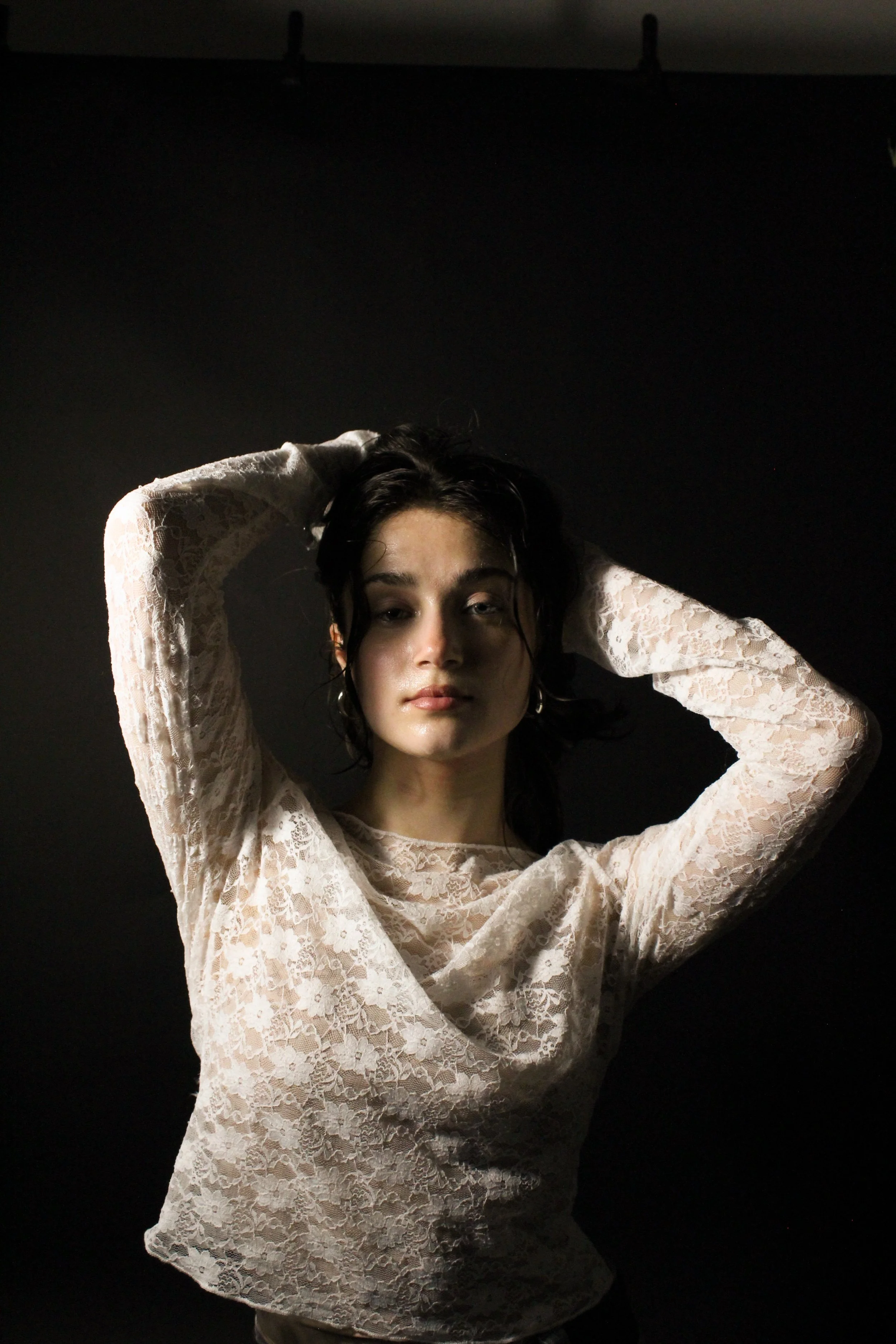 Ein junges Mädchen mit dunklen, nassen Haaren, in einer weißen Spitzenbluse, posiert vor einem schwarzen Hintergrund, mit einer Lichtquelle, die auf ihr Gesicht und Oberkörper fällt.