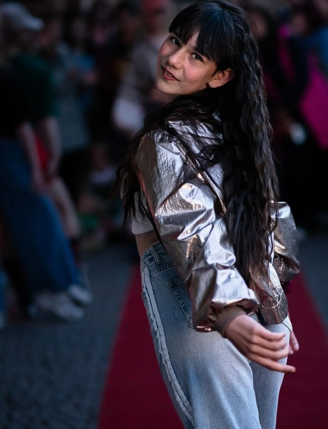 Junge Frau mit langen schwarzen Haaren, silberner Jacke und Jeans posiert in einer Menschenmenge bei Nacht.