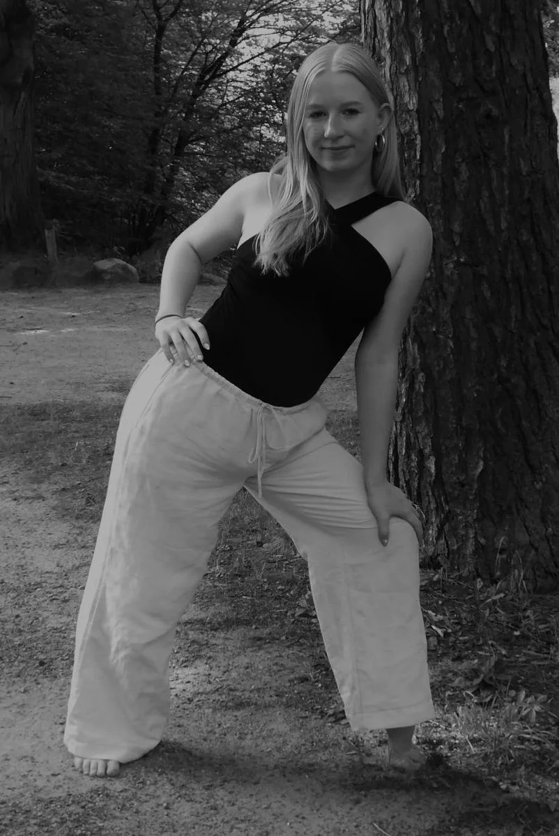 Junge Frau steht im Freien an einem Baum und posiert für ein Foto, trägt eine schwarze Top und weiße Hosen, mit einer Hand an der Hüfte.