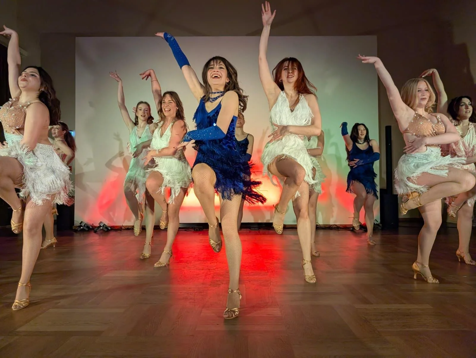 Mehrere Frauen tanzen auf einer Bühne in Tanzkostümen mit Federn, bei einer Performance, mit roten und grünen Lichteffekten im Hintergrund.