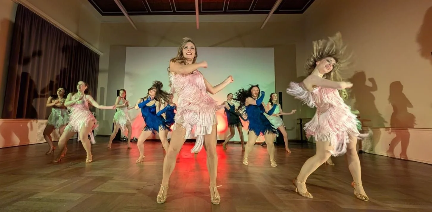 Gruppe von Frauen, die einen Tanz auf einer Bühne aufführen, getragen in bunten Fransen-Kleidern, mit Hemdarten Schuhen, während sie im Scheinwerferlicht tanzen.