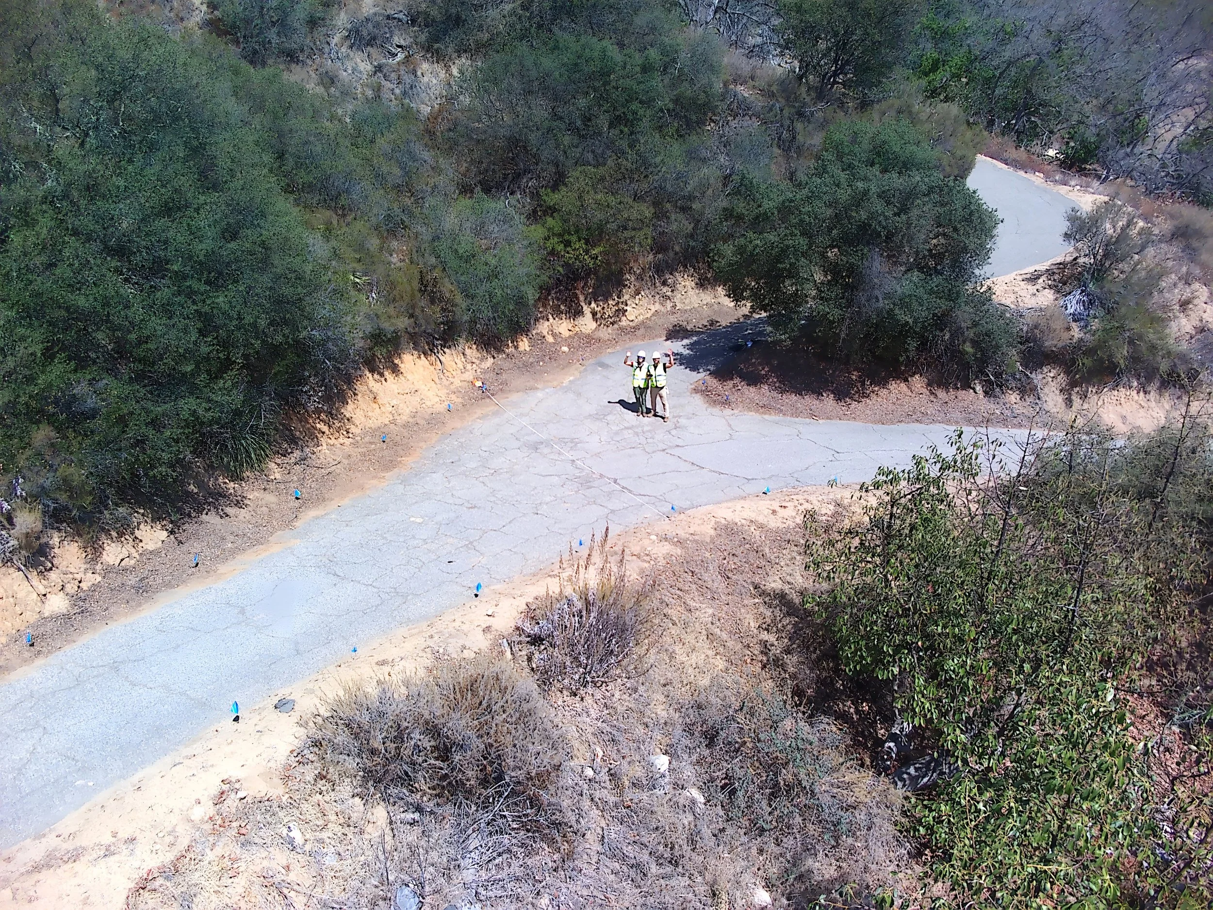 Two workers wearing safety helmets and vests holding tools on a gravel road in a hilly, wooded area.