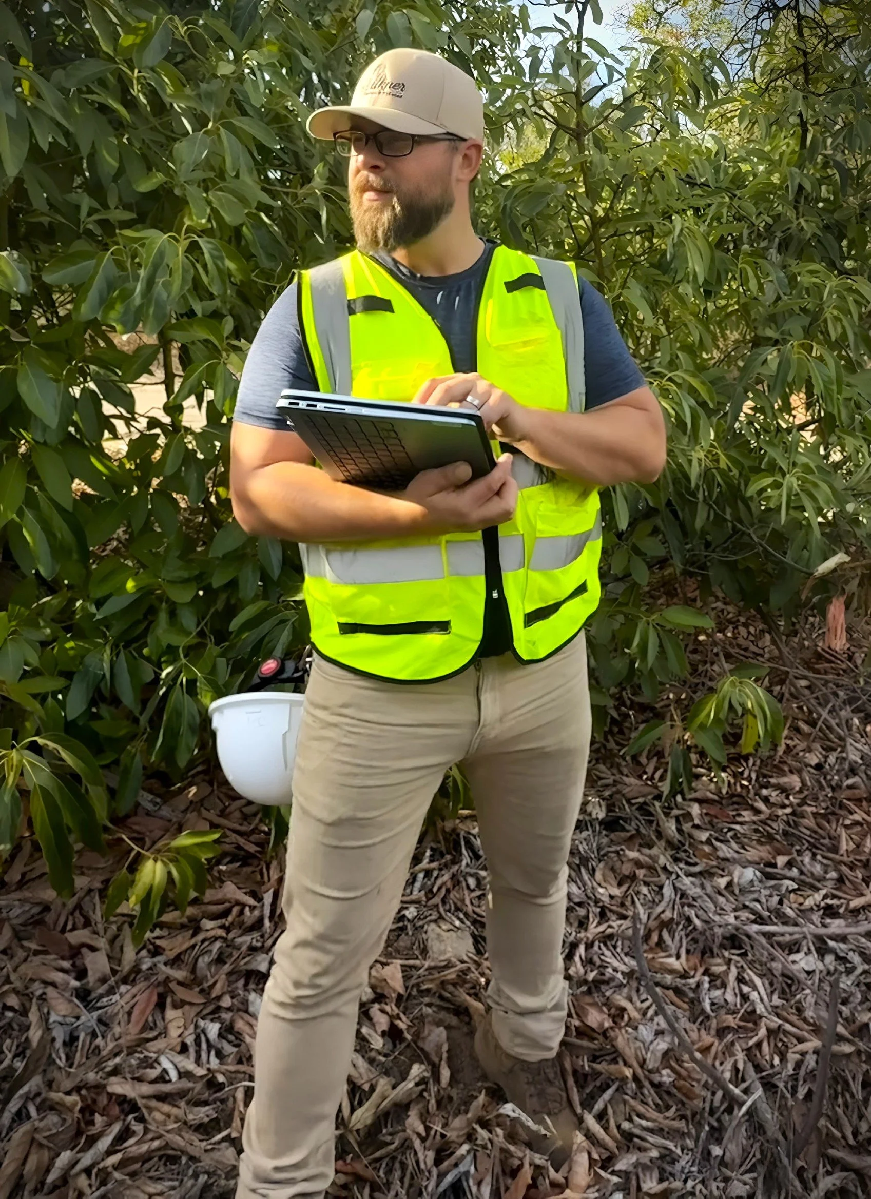 A man wearing a beige hat, glasses, a bright yellow safety vest, and beige pants, holding a laptop and a white bucket, standing outdoors among green leafy trees and fallen leaves.