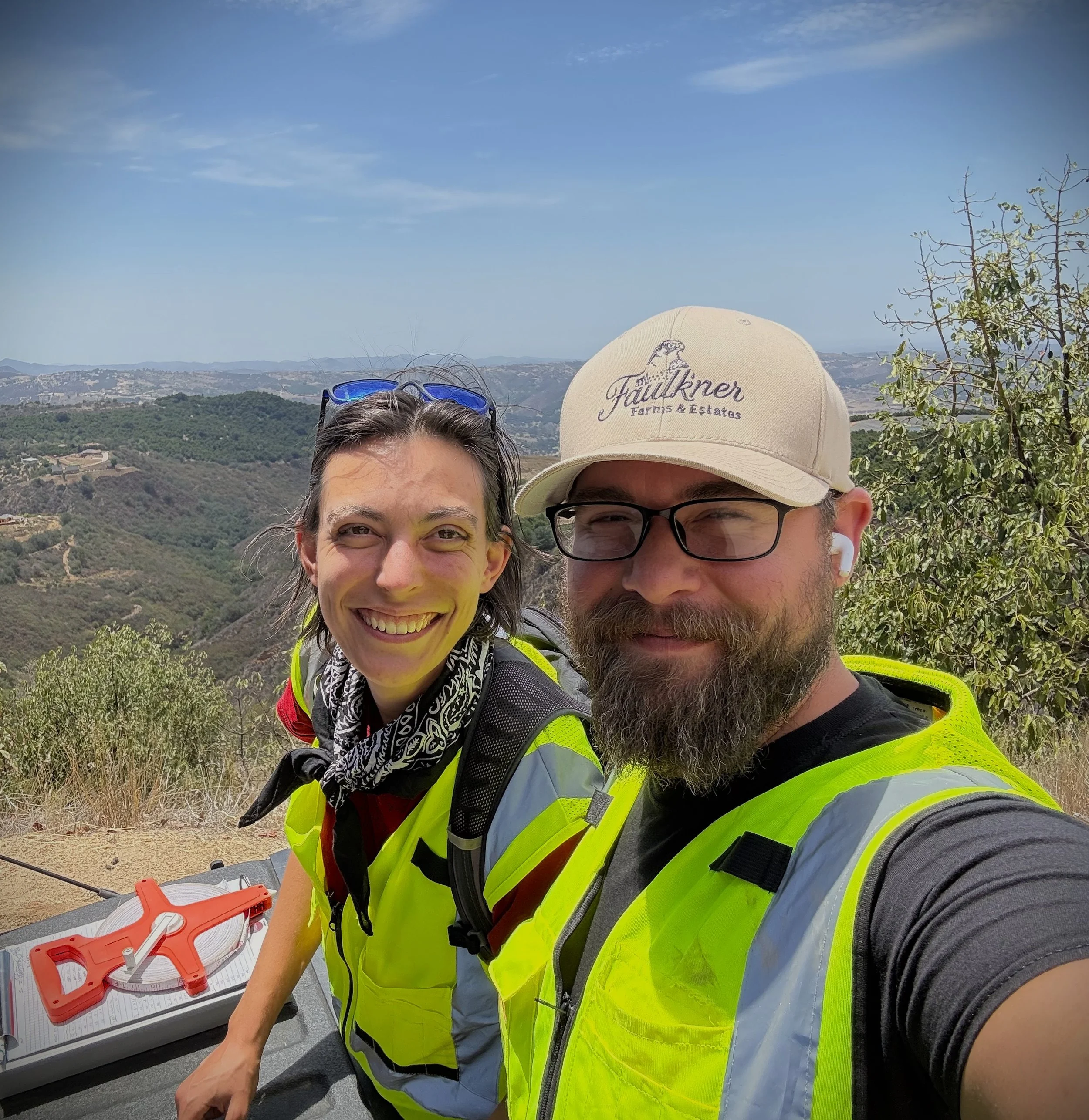 Two people wearing high-visibility safety vests, one with sunglasses on their head and the other with a hat, taking a selfie outdoors with a scenic landscape of rolling hills and blue sky in the background.