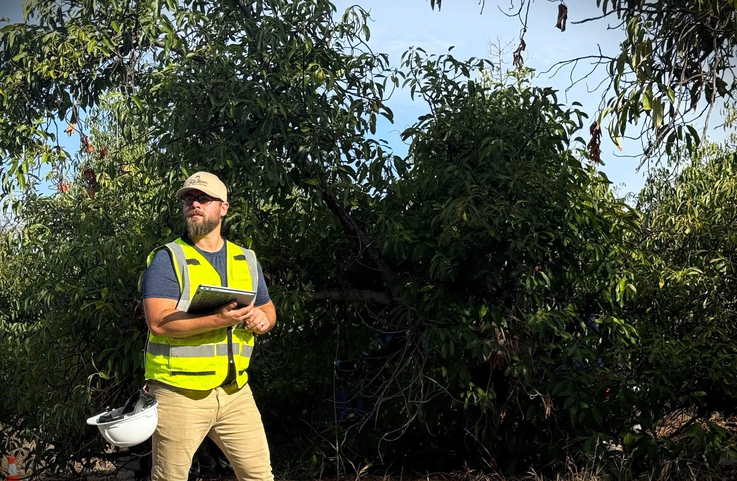 A man wearing a yellow vest and a white helmet, holding a tablet and standing outdoors in front of dense green trees.