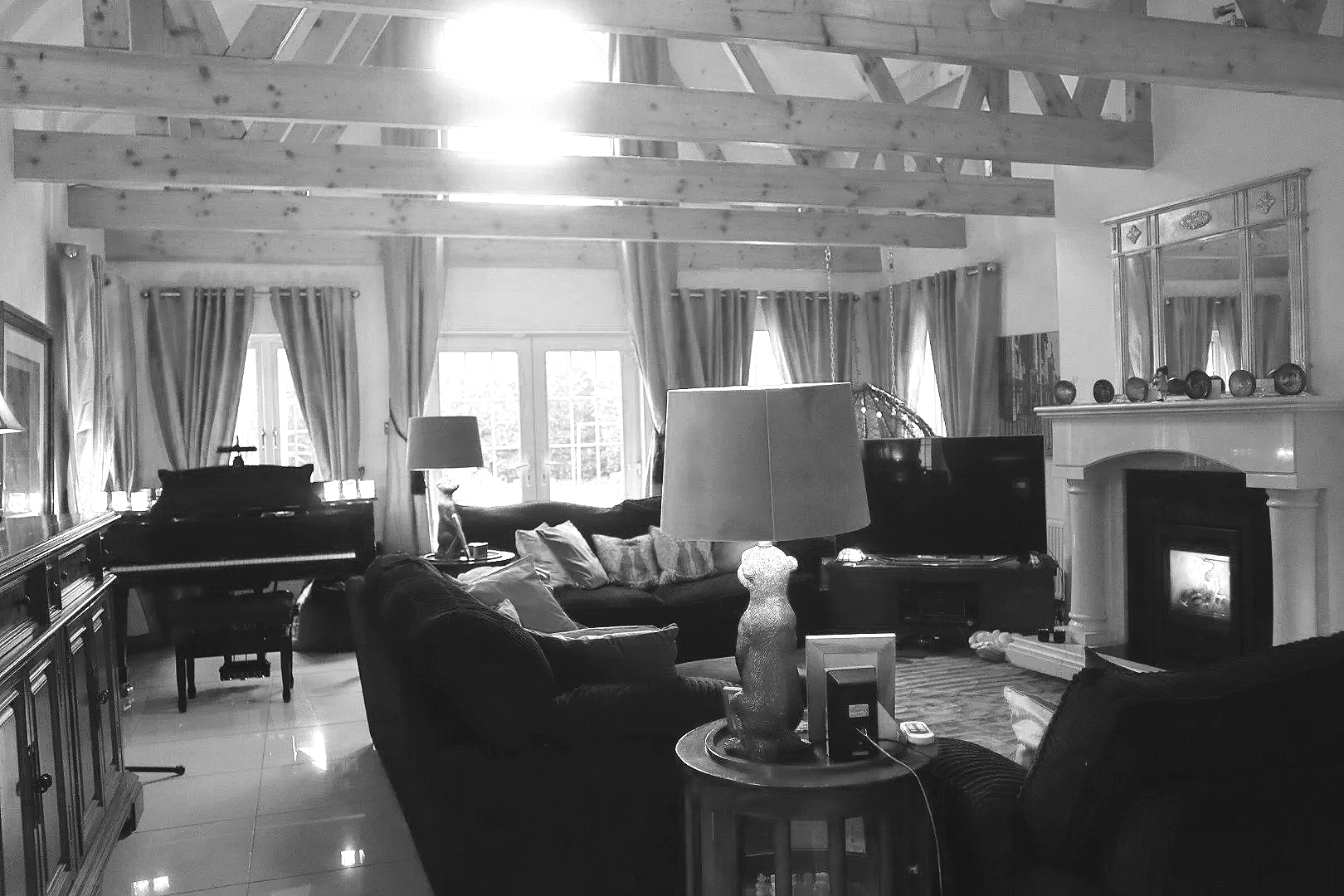 Living room with large windows, curtains, a fireplace, a TV, a piano, and sofas with cushions, decorated with lamps and vases, and exposed wooden ceiling beams.