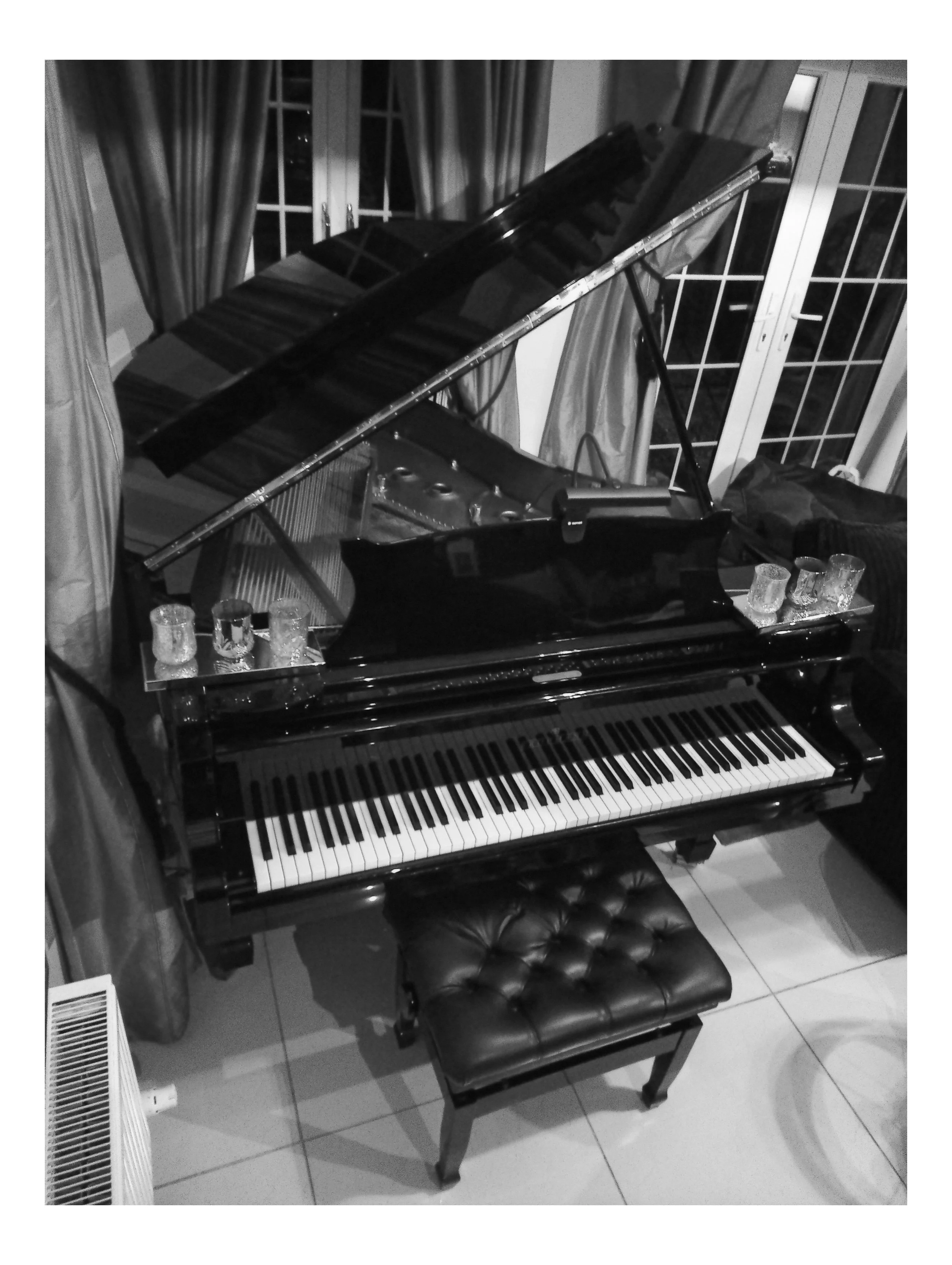 A black grand piano with its lid open, surrounded by glassware, a piano bench in front, and curtains in the background, in a room with tiled floor.