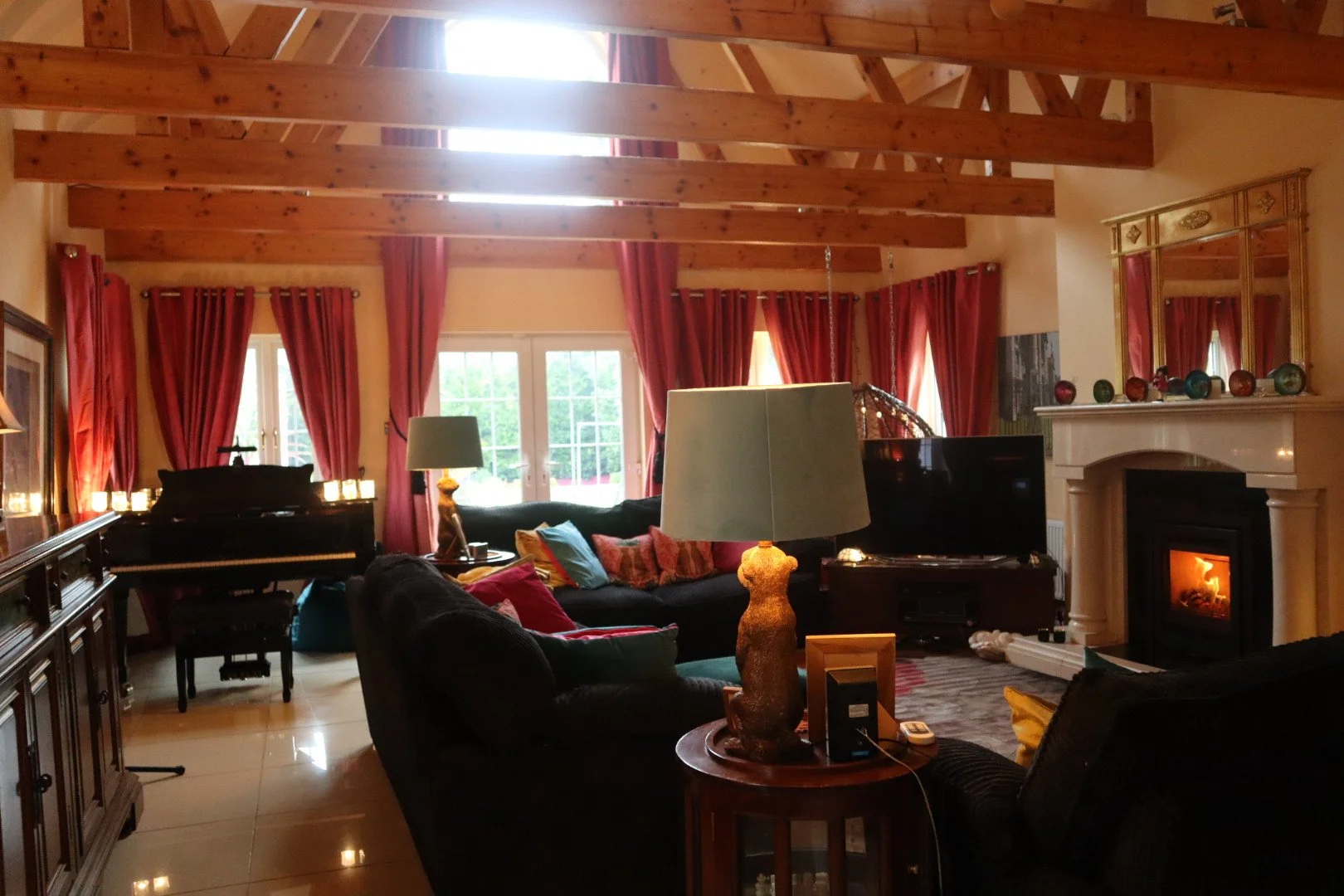 A cozy living room with exposed wooden beams, red curtains, a fireplace with a mantel decorated with colorful ornaments, a black sofa with pillows, a TV, a piano, and lamps, with sunlight coming through large windows.