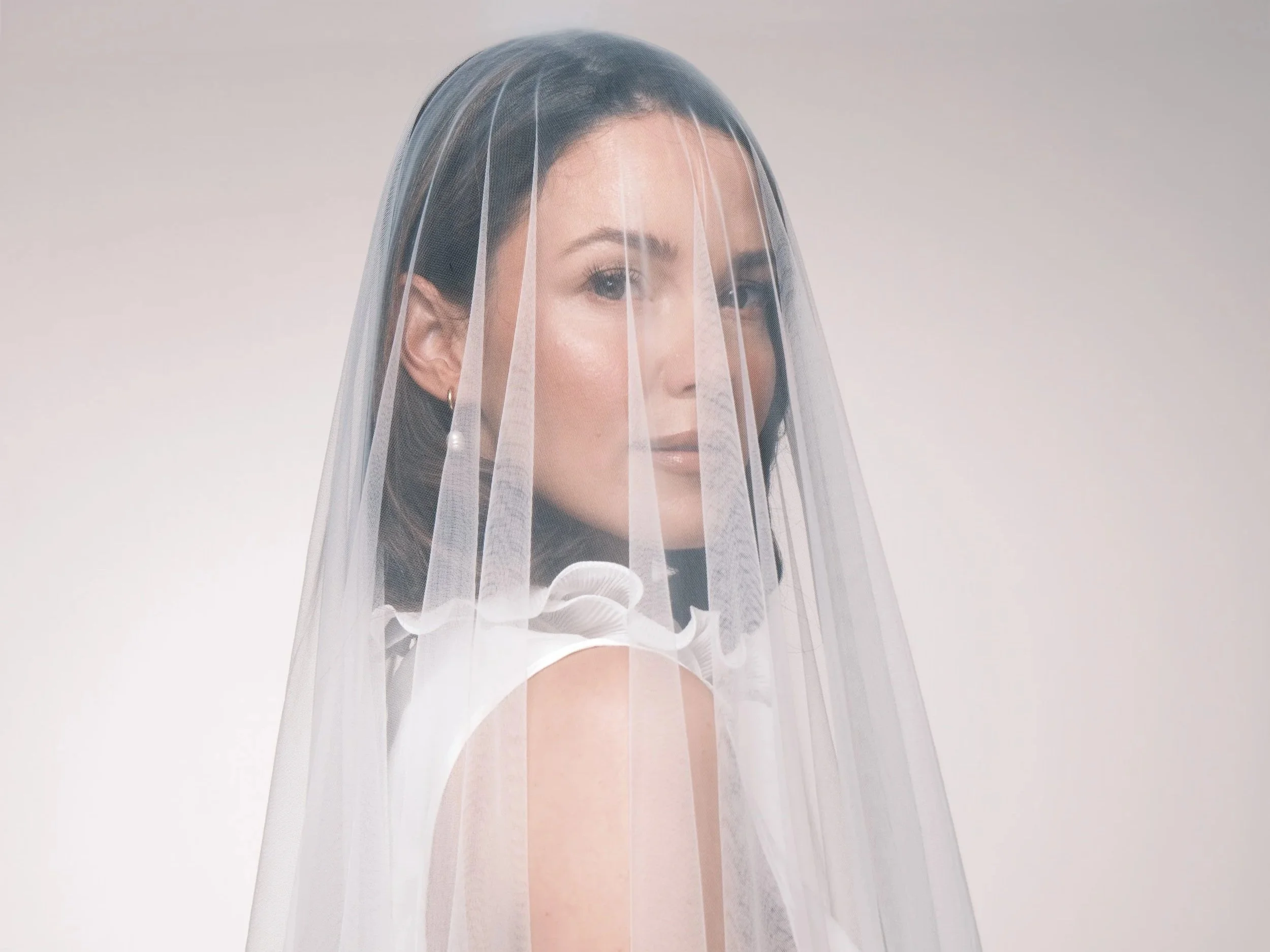 Bride under white veil with brunette curled bob looking directly at camera on wedding day