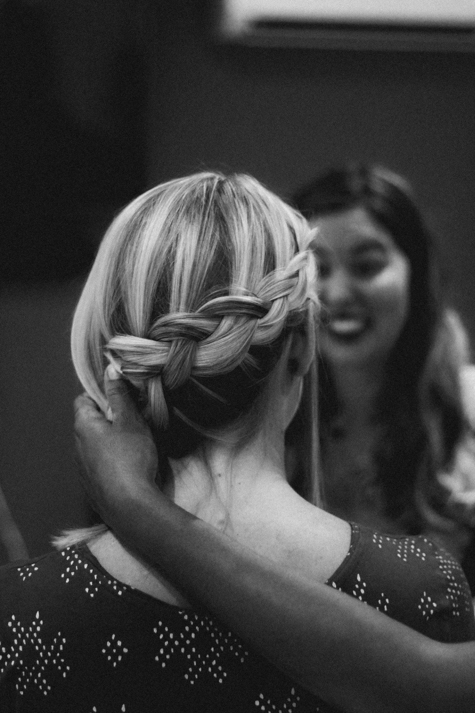A woman with blonde hair in a braid is adjusting it while another woman with dark hair looks on, smiling, in the background. The image is in black and white.