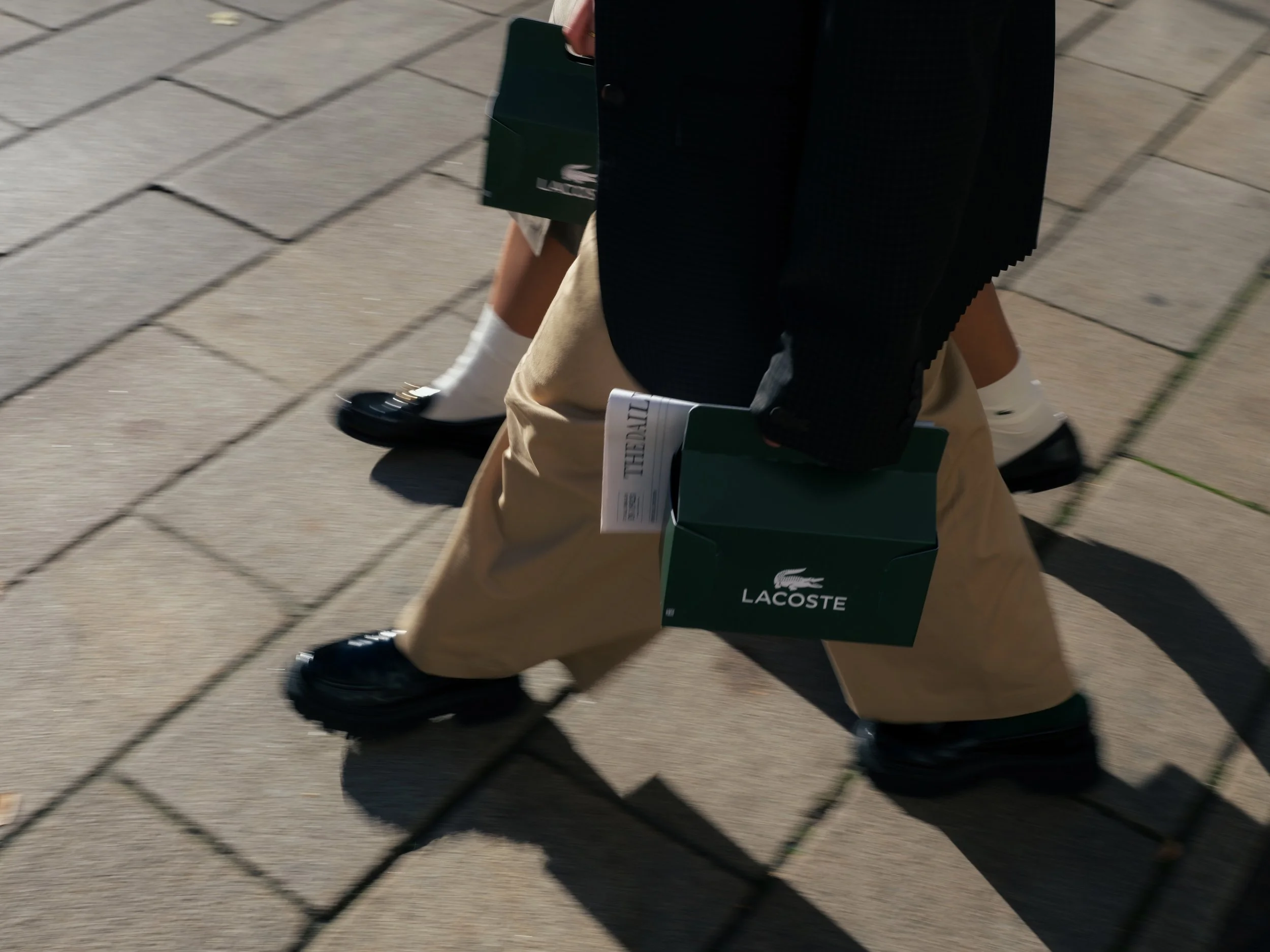 Person, die Laufschuhe trägt und braune Hosen sowie einen schwarzen Blazer trägt, läuft auf einem Bürgersteig und hält grüne Lacoste-Taschen.