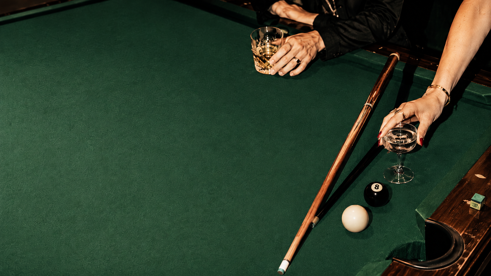 Nahaufnahme eines Billardtisches mit Spielerinnen, die ein Glas Wasser halten, und Billardkugeln, darunter die 8 und die weiße Kugel, sowie eine Queue.