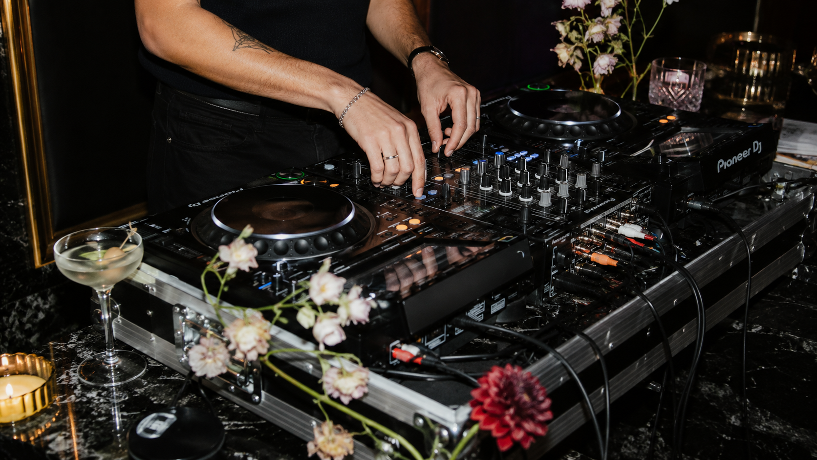 Ein DJ, der an einem Mischpult auflegt, umgeben von Blumen und dekorativen Gegenständen in einem dunklen Raum.