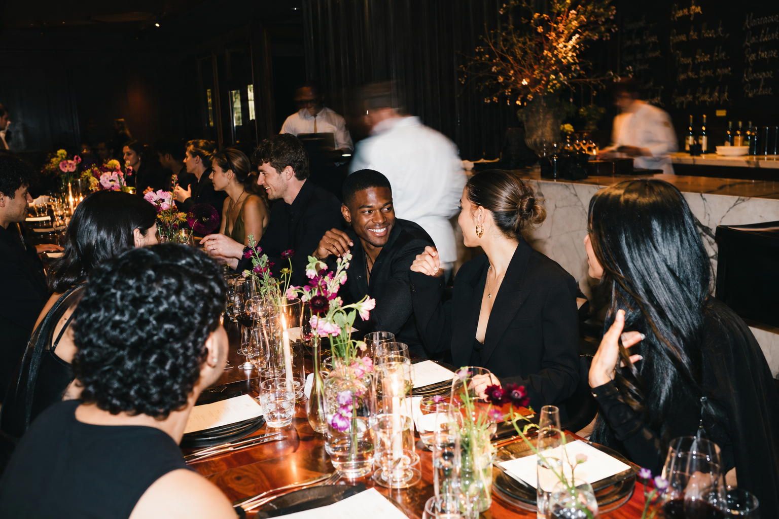 Menschen bei einem festlichen Dinner, unterhalten sich und lachen in einem eleganten, dunklen Raum mit Blumen und Kerzen auf dem Tisch.
