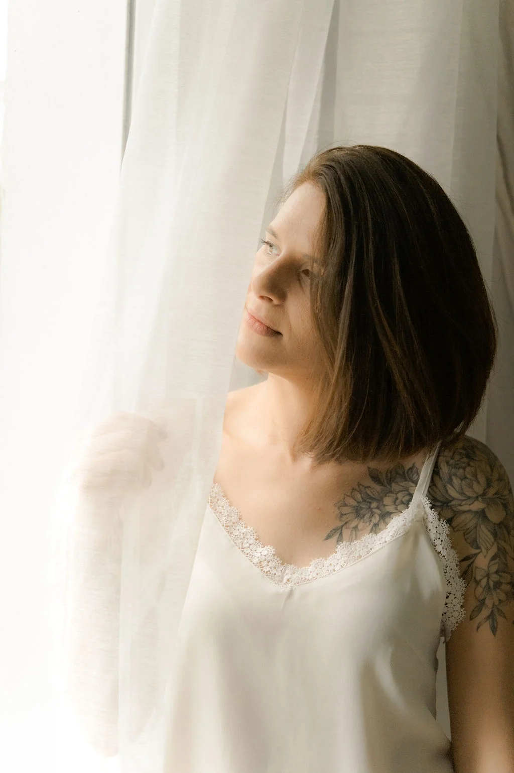 Portrait de Charlotte GANDON, vidéaste et photographe en Savoie. La photographie a été prise en studio en lumière naturelle. Charlotte regarde par une fenêtre avec un voilage qu'elle touche de sa main droite.