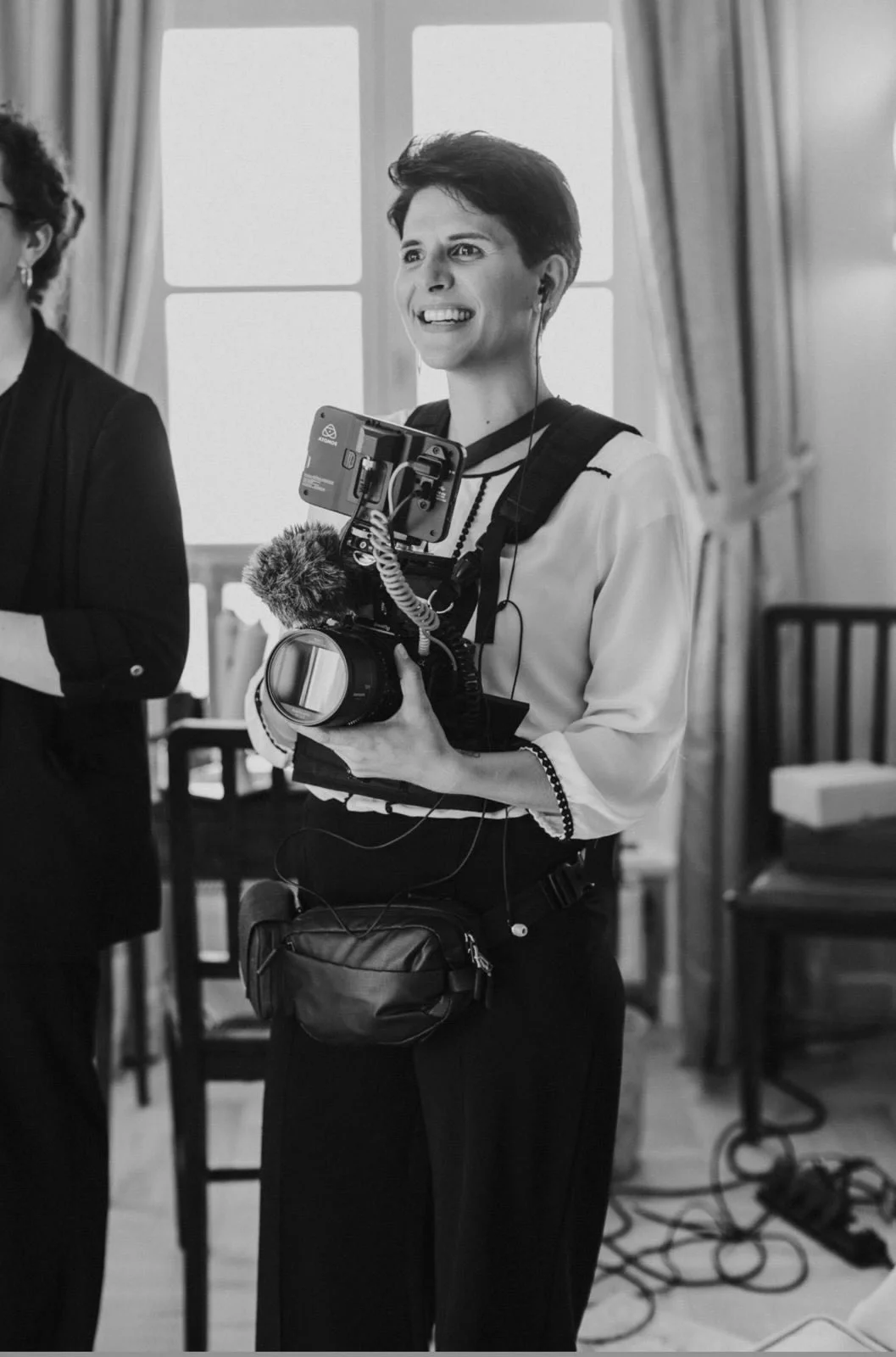 Portrait de Charlotte GANDON, vidéaste de mariage en Savoie. La photo est en noir et blanc et y voit Charlotte caméra à la main entrain de sourire en regardant la mariée.