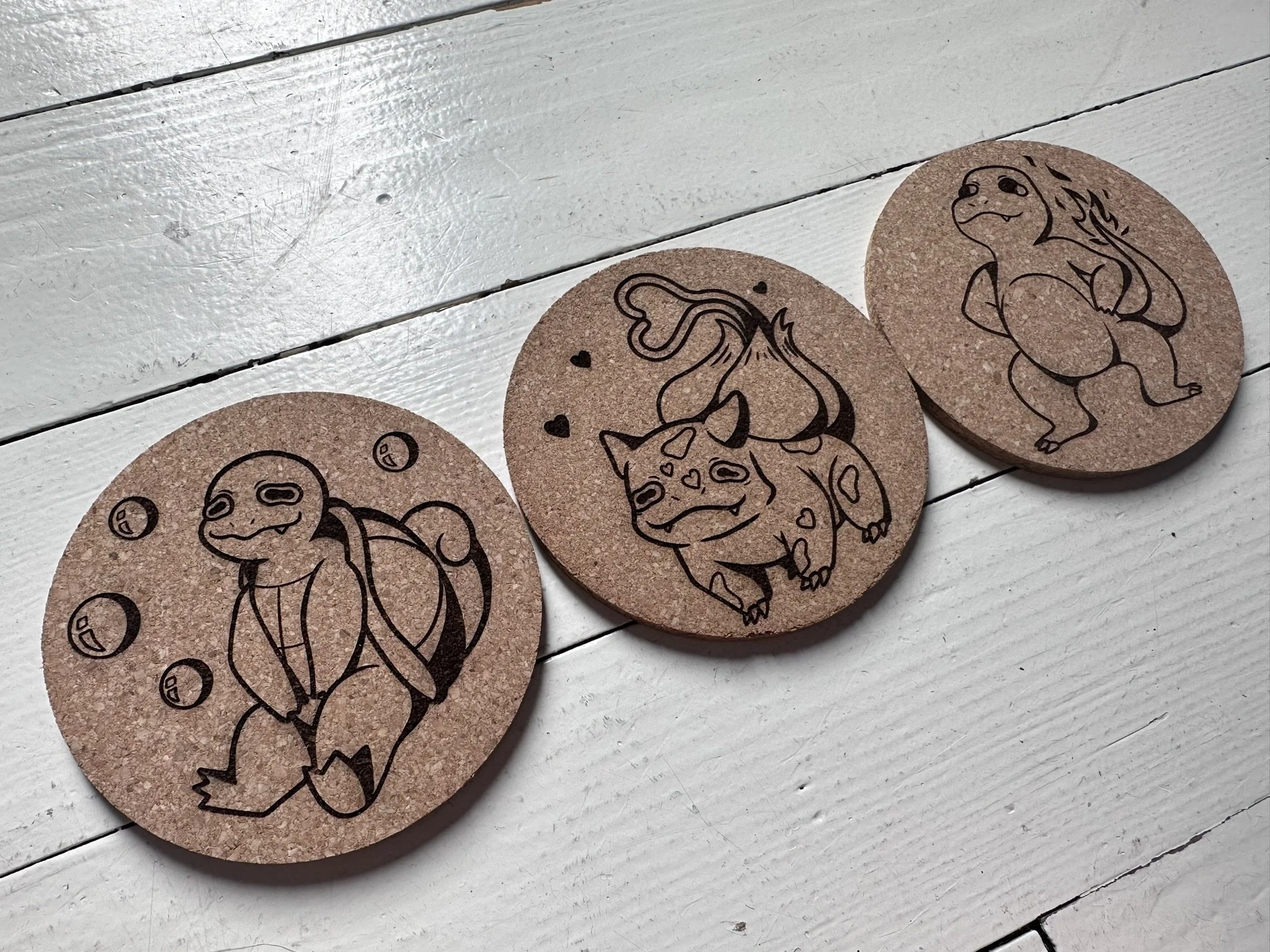 Set of three round cork coasters, each with a line drawing of a Pokémon character: Squirtle, Bulbasaur, and Charmander.