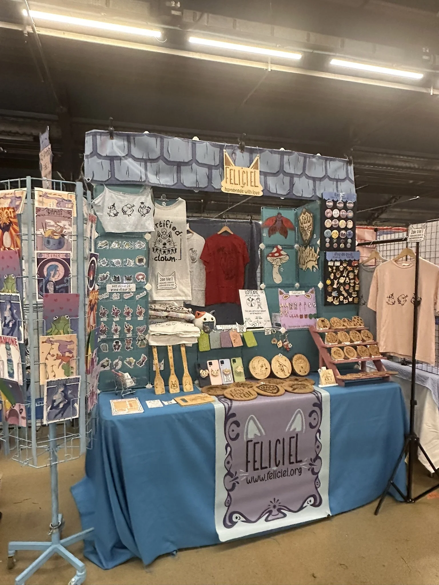 A booth at a market with various art and merchandise, including stickers, buttons, t-shirts, wooden items, and postcards, all promoting Feliciel, a handmade brand. The booth has a blue tablecloth and a sign with the Feliciel logo and website.