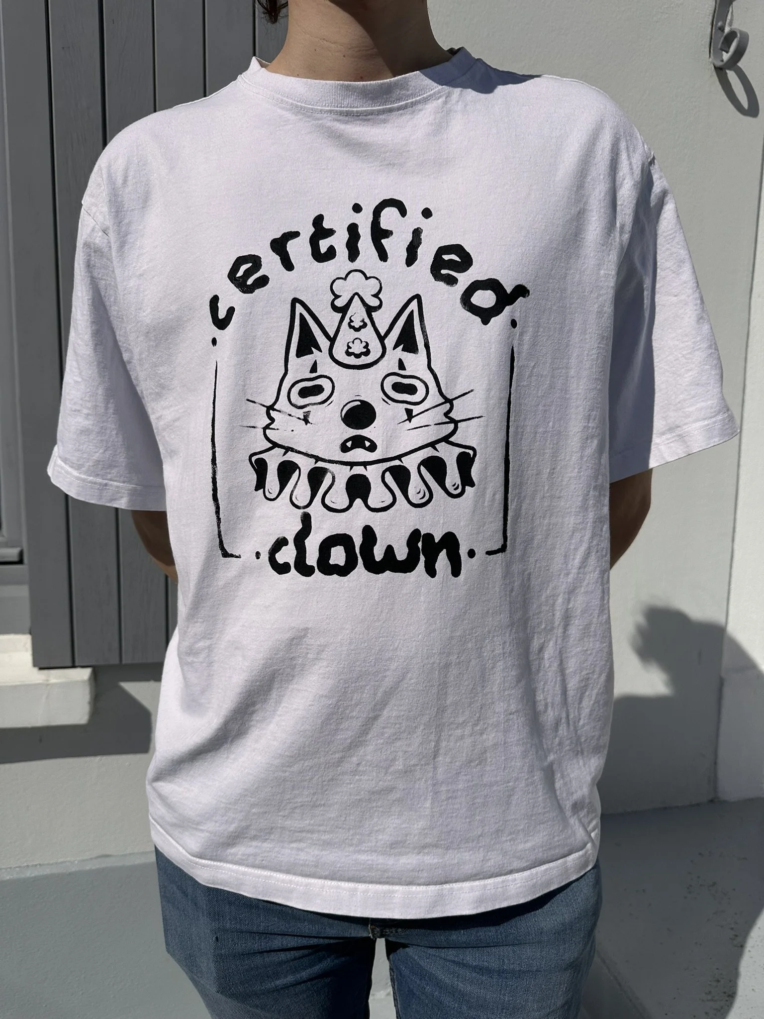 White T-shirt worn by a person, featuring a black line drawing of a cat with a unicorn horn and a ruffled collar, with the words 'certified clown' printed above and below the drawing.