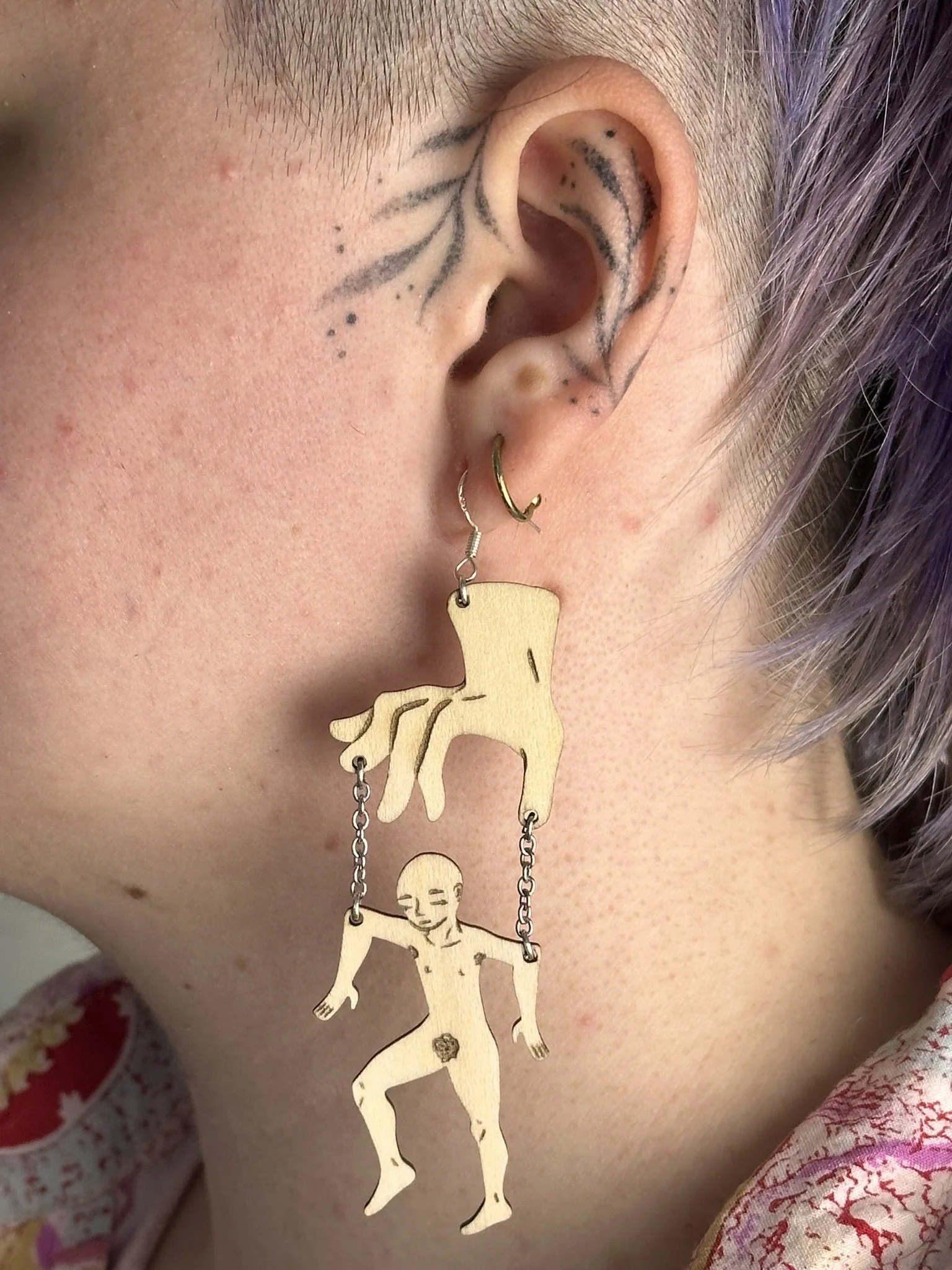 Close-up of a person's ear with multiple earrings including a gold hoop, a gold cartilage ring, and a large dangling earring with cut-out figures of a hand and a humanoid figure connected by chains, showing a puppet and it's puppeteer.