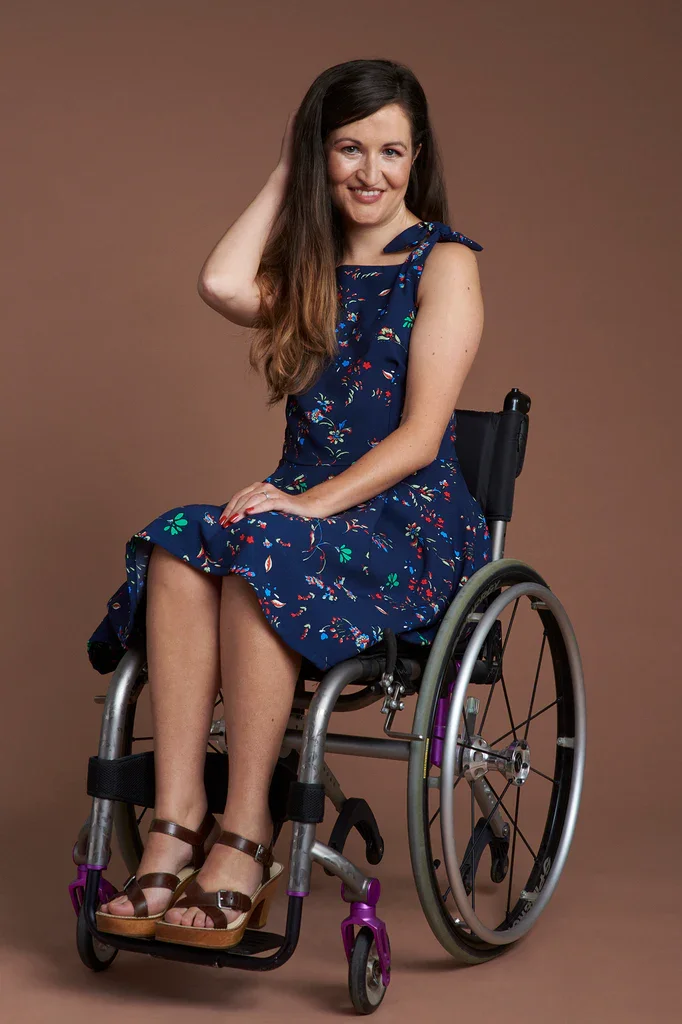 Marie Lawlor is wearing a blue summer dress and sandals and is smiling. She is using a wheelchair