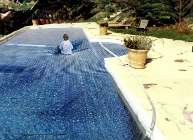 Enfant assis sur un filet de sécurité couvrant une piscine, entourée de plantes en pot et de transats.