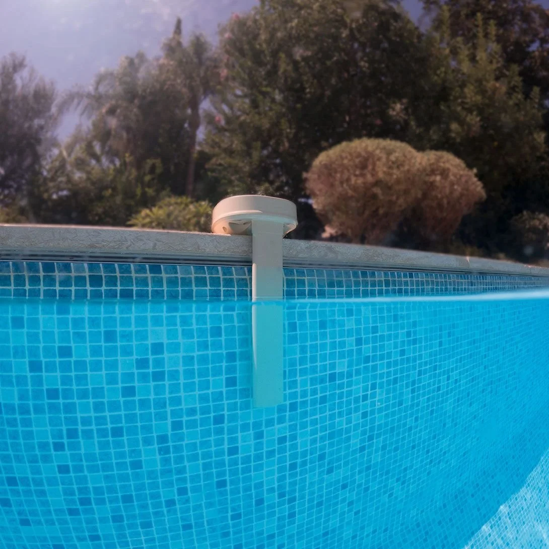 Capteur ou dispositif de sécurité fixé sur le bord d'une piscine avec un carrelage bleu, et des arbres en arrière-plan.