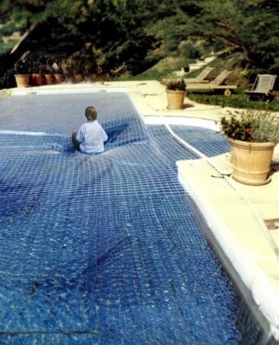 Un enfant assis sur une couverture de piscine bleue, entouré de jardinières et de chaises longues dans un jardin verdoyant.