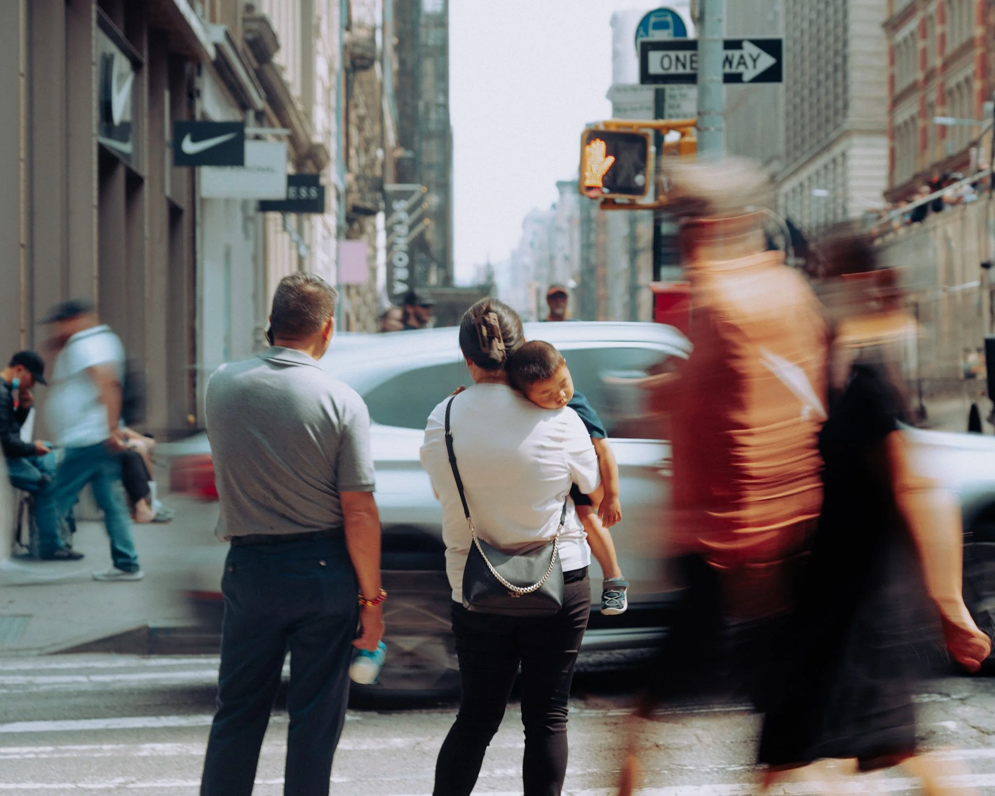 A Mother holds her sleeping child while standing on the corner of a busy New York City street