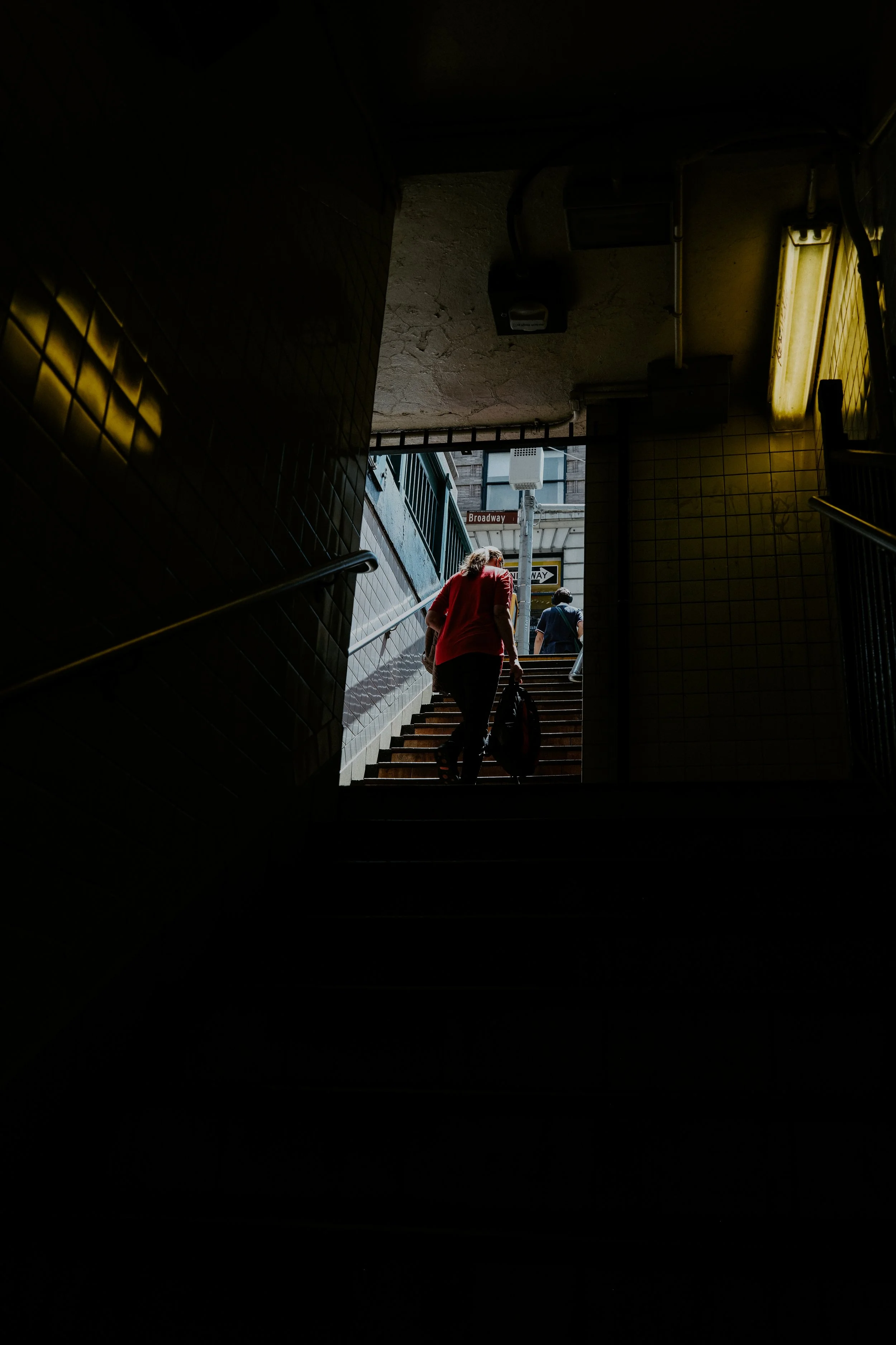 A woman leaves the shadowy environment of a New York City subway and climbs the steps towards the light. The yellow light adds to the ambience of the dimly lit subway station.