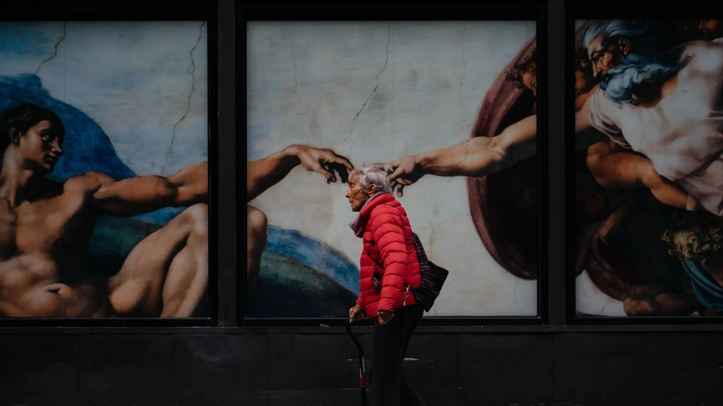 An elderly woman in a red jacket walking past a large mural of Michelangelo's The Creation of Adam on a New York City street.