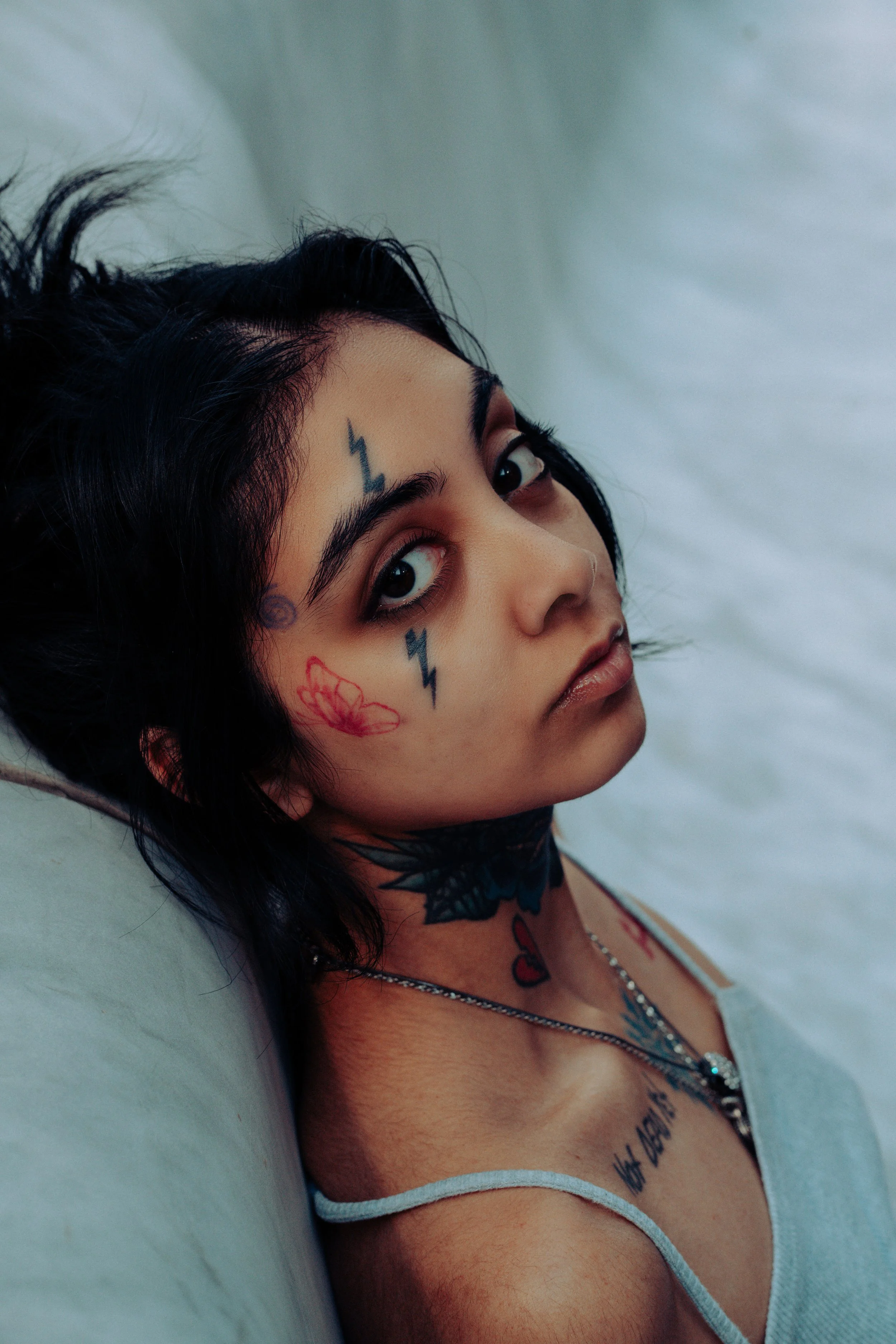 Close-up of a woman with black hair in an urban enviornment, showing various tattoos on her face and chest, including lightning bolts and butterfly designs, with dark eye makeup and a silver necklace.