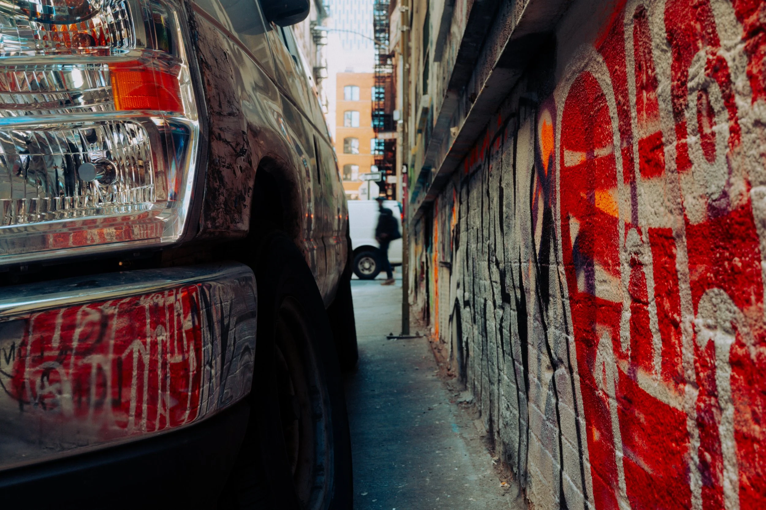A close-up of the front of a black vehicle parked along a colorful, graffiti city street. In the background, there is a person walking and buildings with windows.