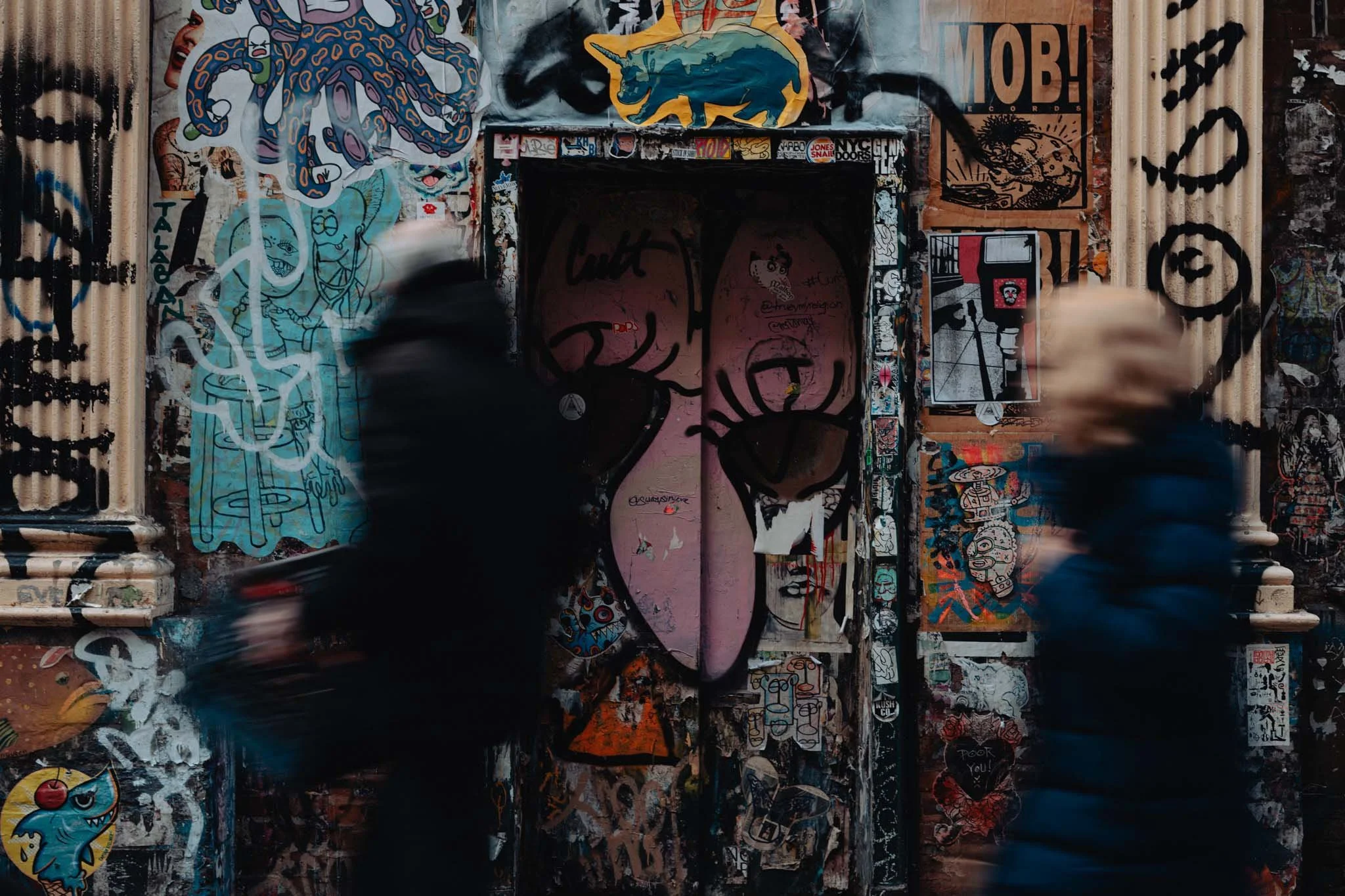 Two people hurridley pass the screen in front of a graffiti covered building.