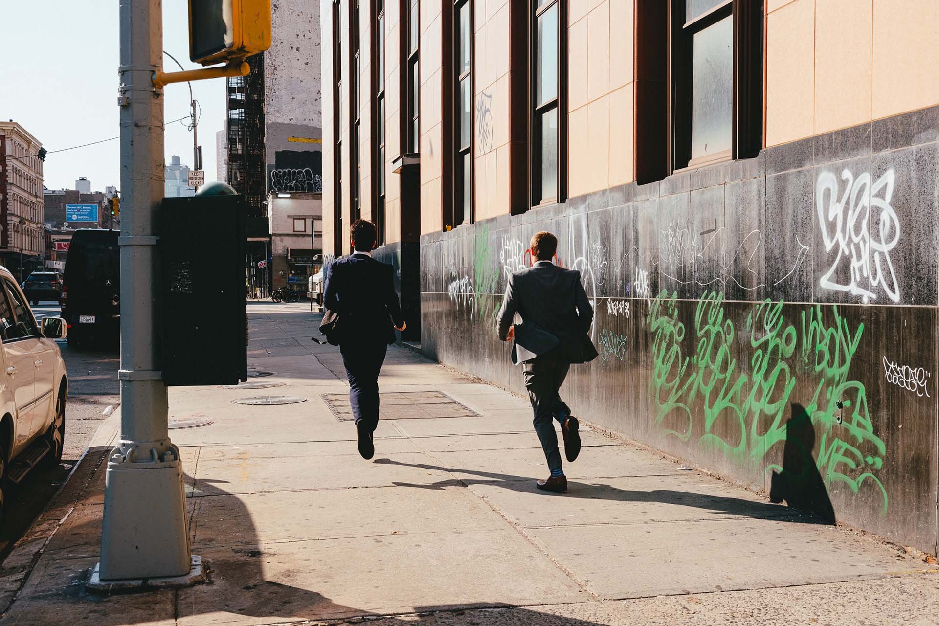 In this photo, two suited men sprint away from the camera, their backs turned towards us as they pass by a graffiti-covered wall.