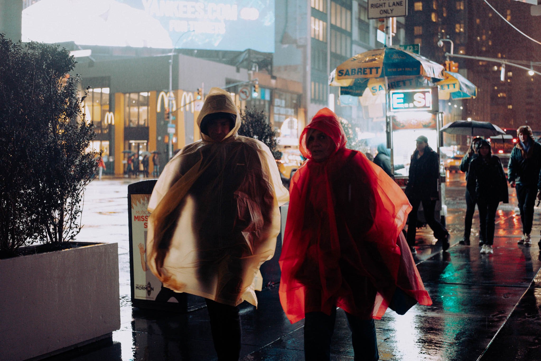 Two people, one in a red poncho and the other in a yellow poncho, hurriedly walk through the rain in Times Square, New York