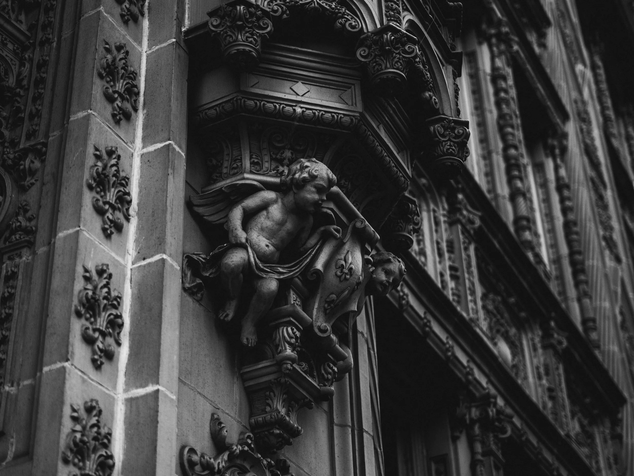 Black and white photo of an ornate building facade with detailed sculptures and carvings, including a cherub holding a shield.