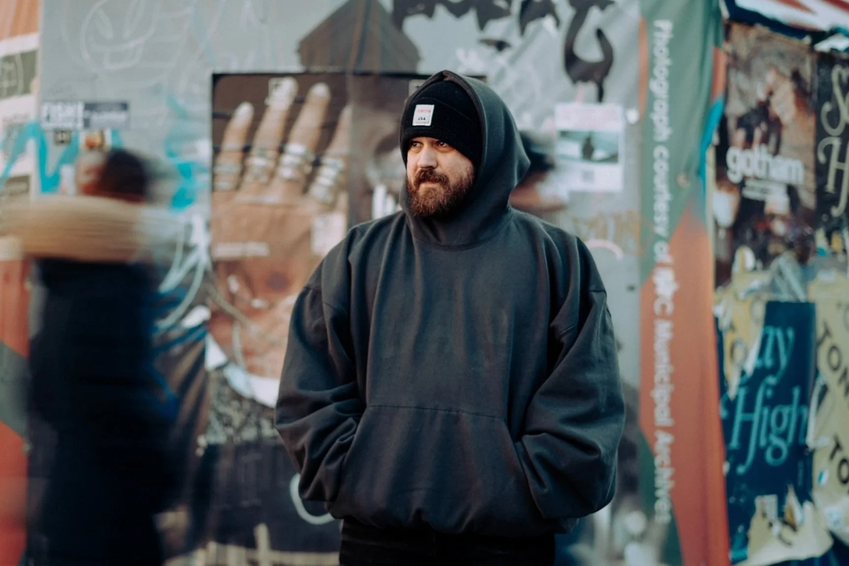 A man wearing a black hoodie and beanie stands in front of a colorful, graffiti-covered wall, looking to his right with a serious expression.